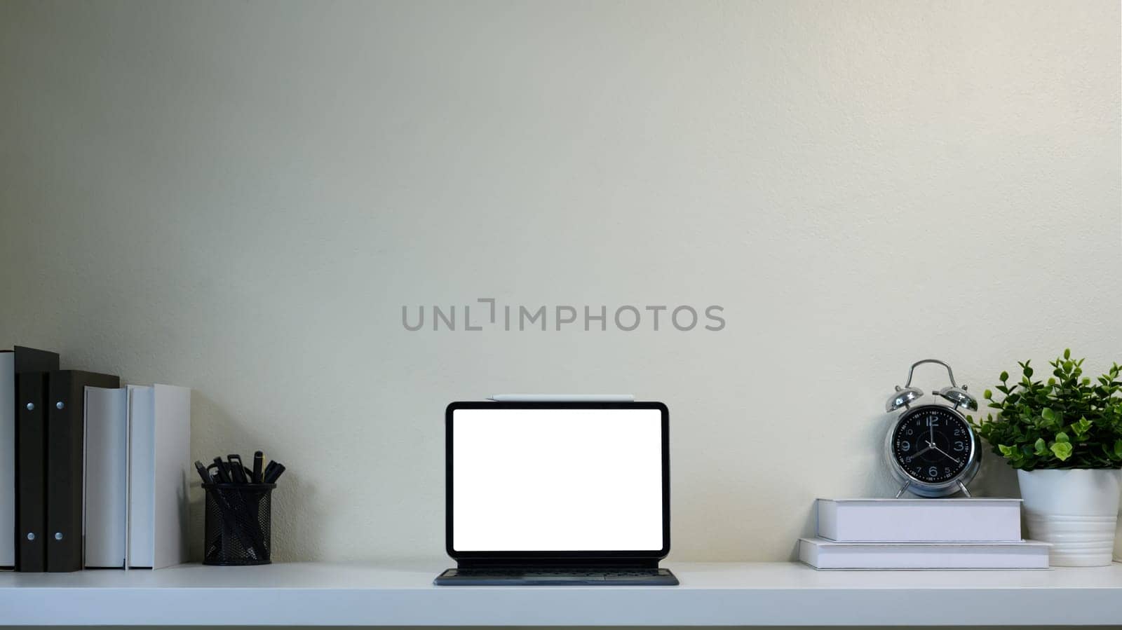 Modern workspace with digital tablet on white desk, surrounded by books, clock and potted plant.