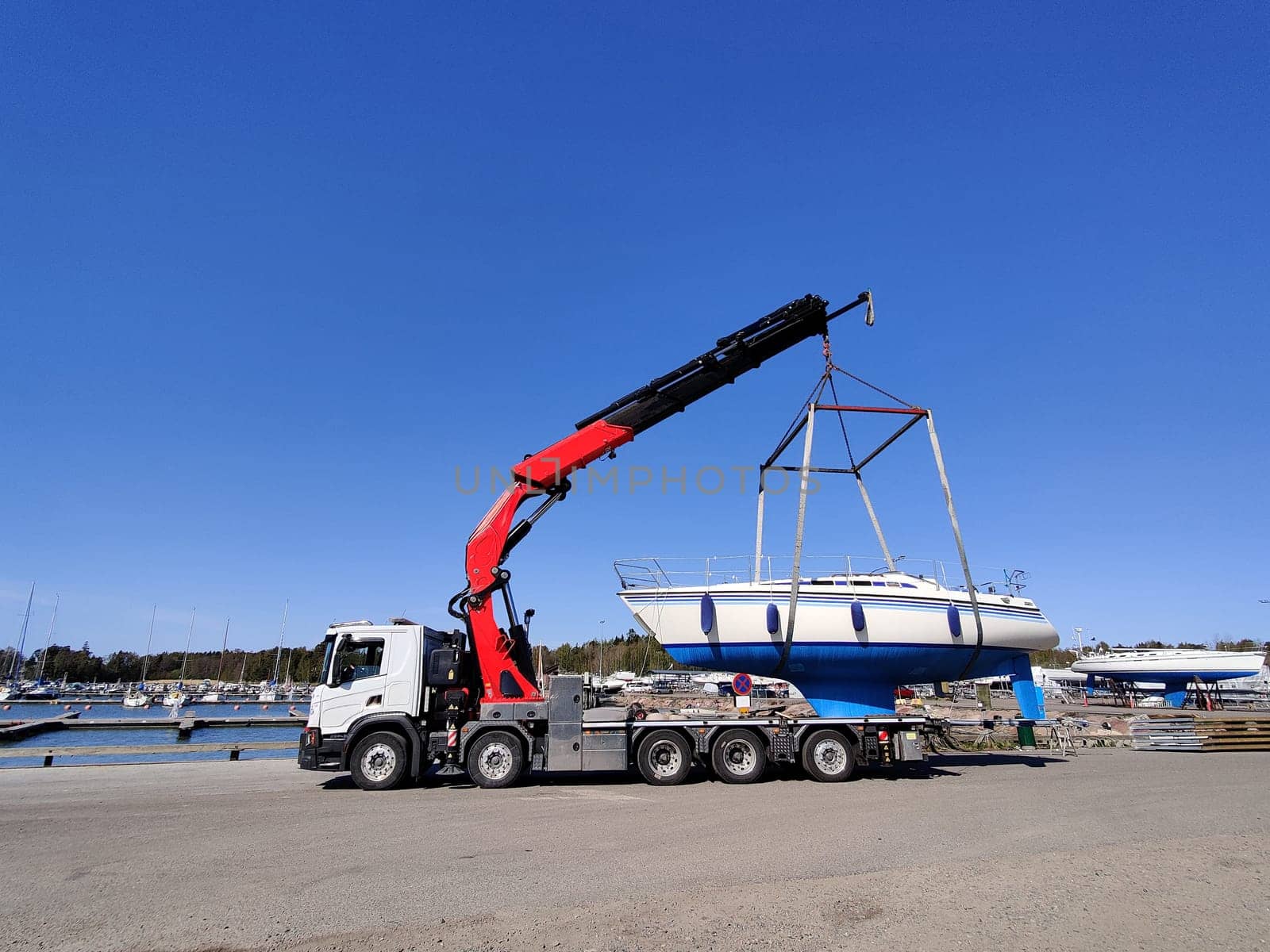A sailboat lifted by a crane in Suomenoja harbor in Espoo, Finland. A man is holding a rope, guiding the boat as it is suspended in the air by lifting straps