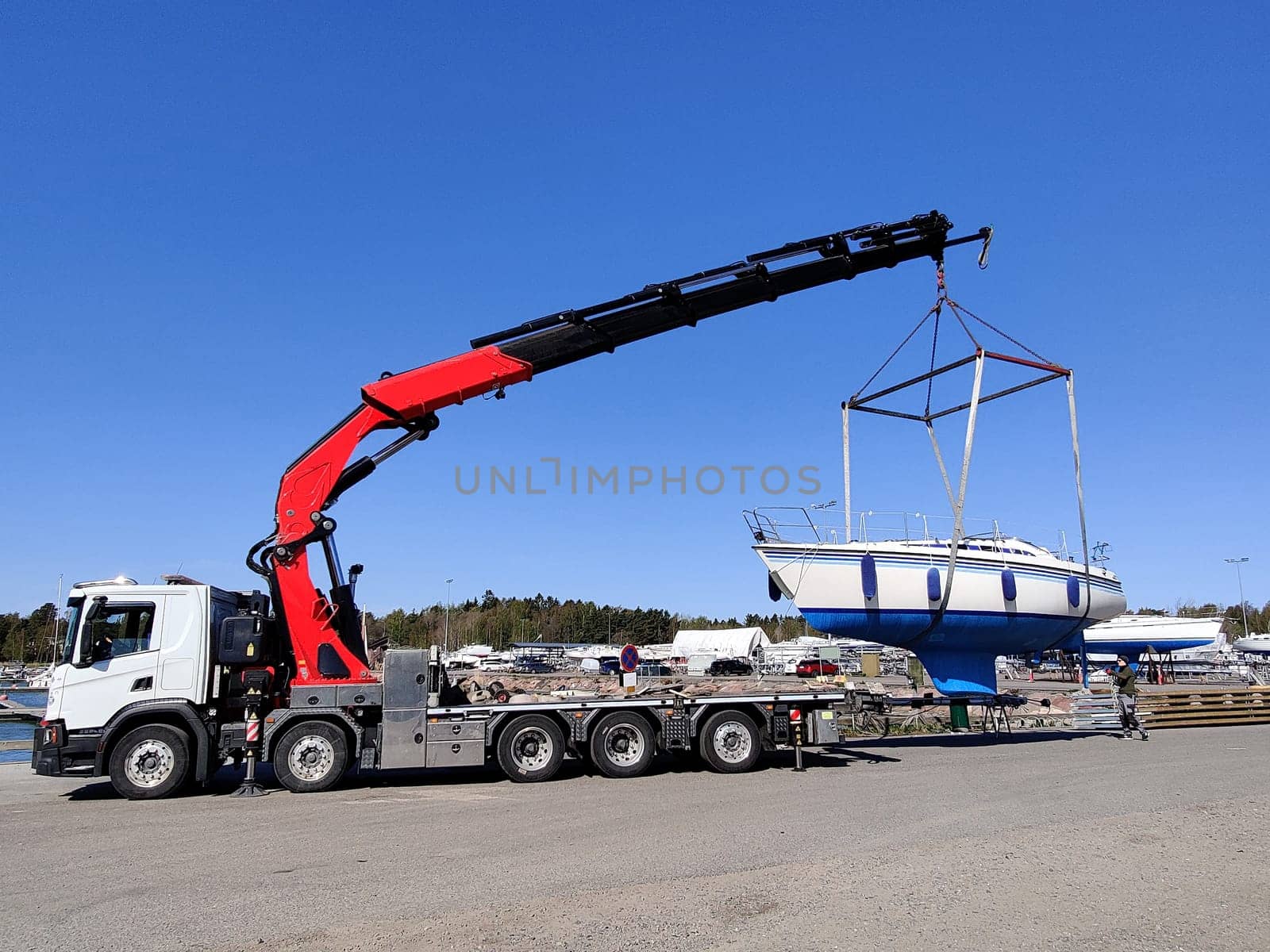 A sailboat lifted by a crane in Suomenoja harbor in Espoo, Finland. A man is holding a rope, guiding the boat as it is suspended in the air by lifting straps
