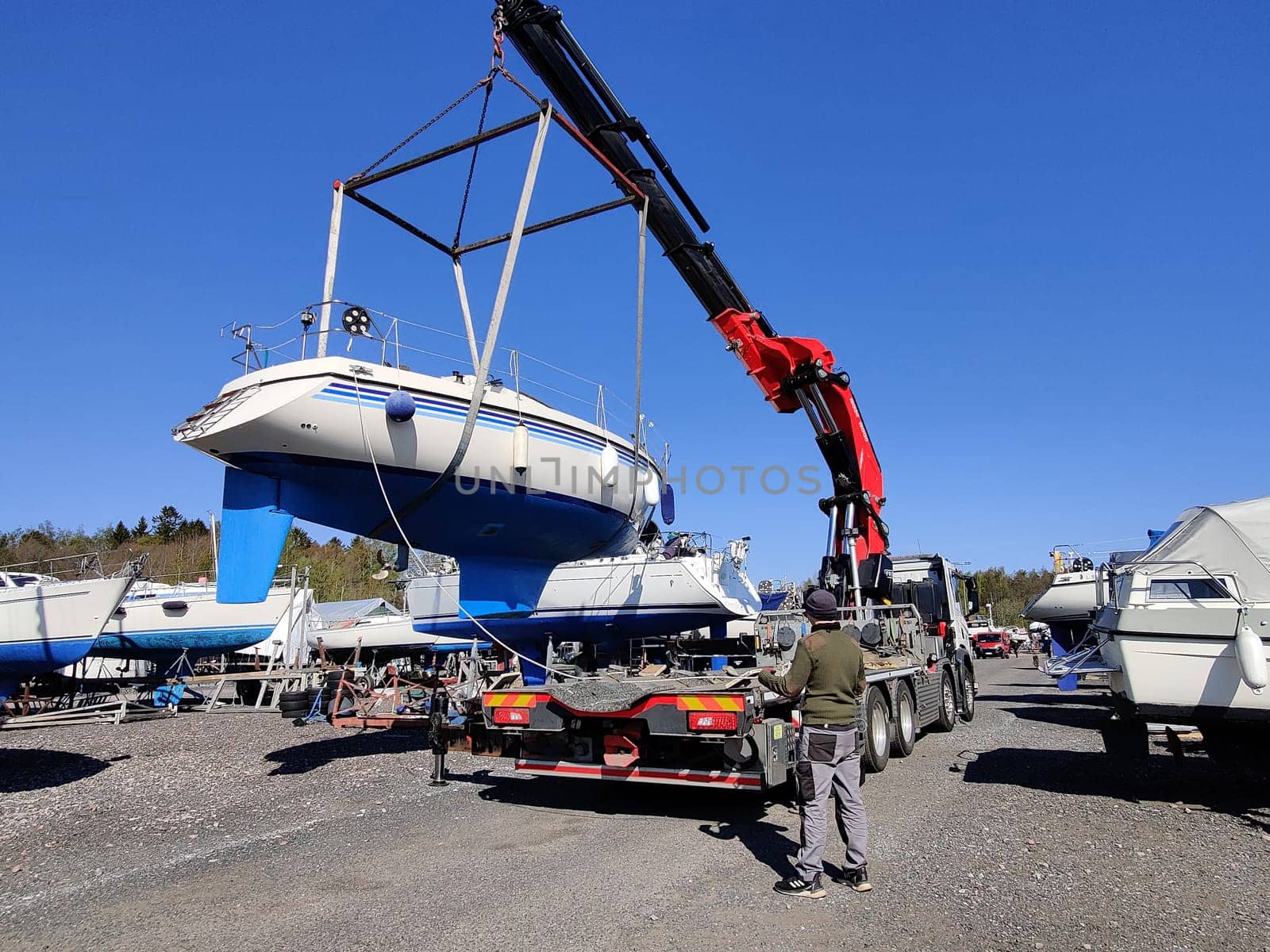 A sailboat lifted by a crane in Suomenoja harbor in Espoo, Finland. A man is holding a rope, guiding the boat as it is suspended in the air by lifting straps