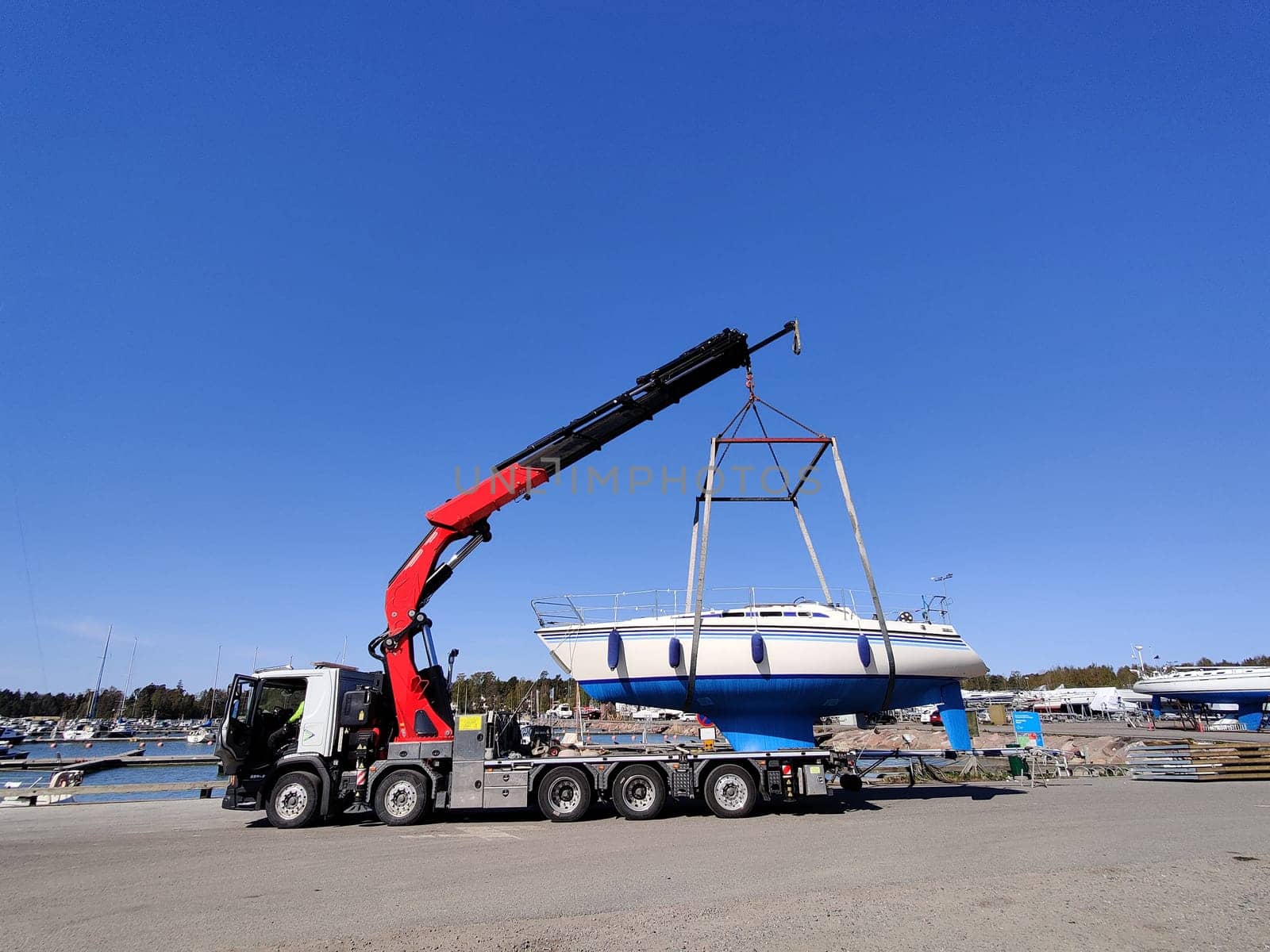 A sailboat is being lifted by a crane in a harbor in Suomenoja, Espoo, Finland. The vessel suspended in the air by heavy lifting straps as it is moved for maintenance or seasonal storage