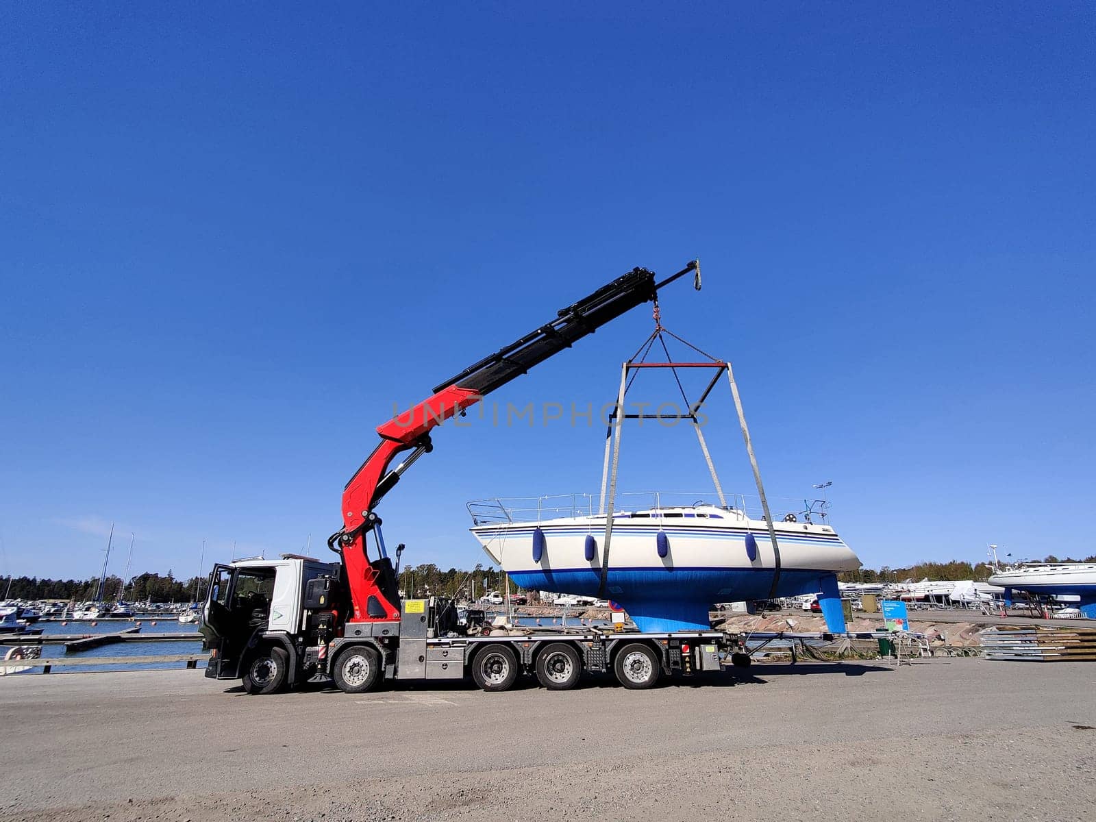 A sailboat is being lifted by a crane in a harbor in Suomenoja, Espoo, Finland. The vessel suspended in the air by heavy lifting straps as it is moved for maintenance or seasonal storage