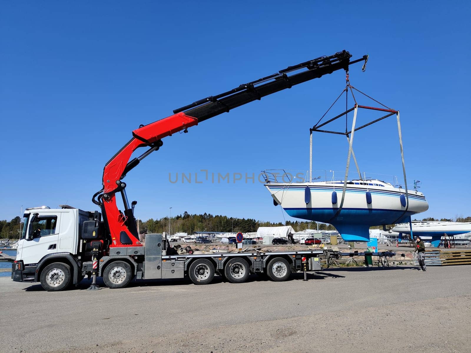 A sailboat lifted by a crane in Suomenoja harbor in Espoo, Finland. A man is holding a rope, guiding the boat as it is suspended in the air by lifting straps