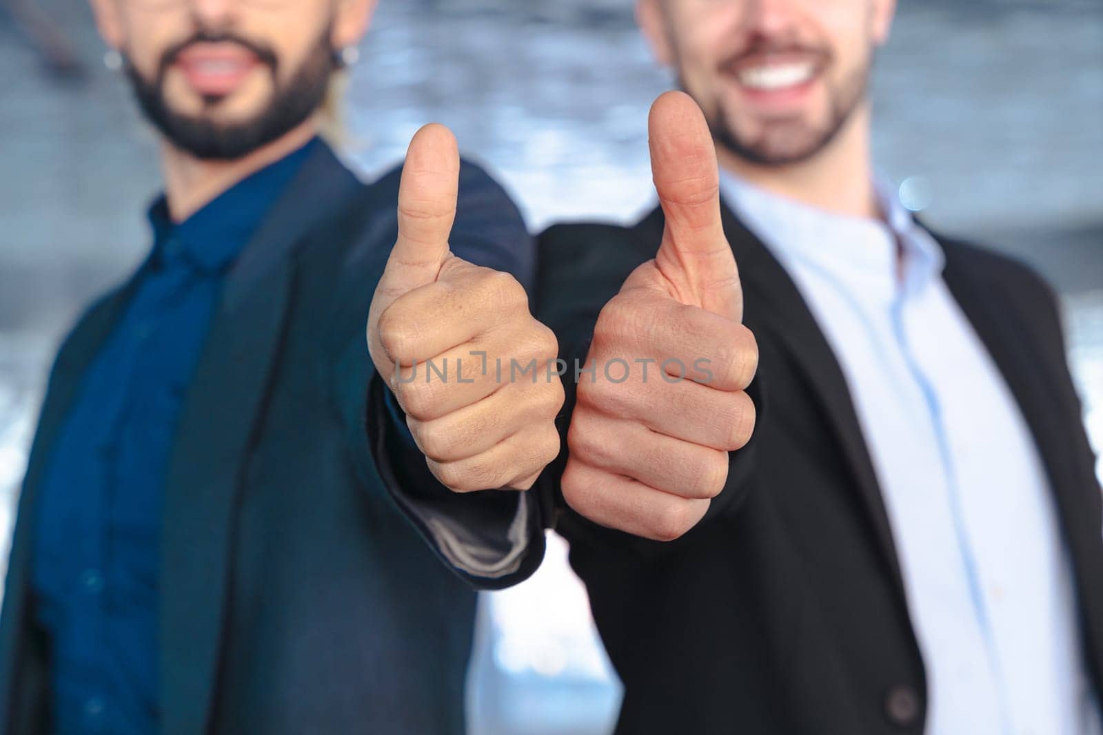 Unrecognizable Two young businessmen in formal suits smiling confidently at the camera, giving thumbs up gesture inside a modern glass office with reflections and stylish interior design elements.