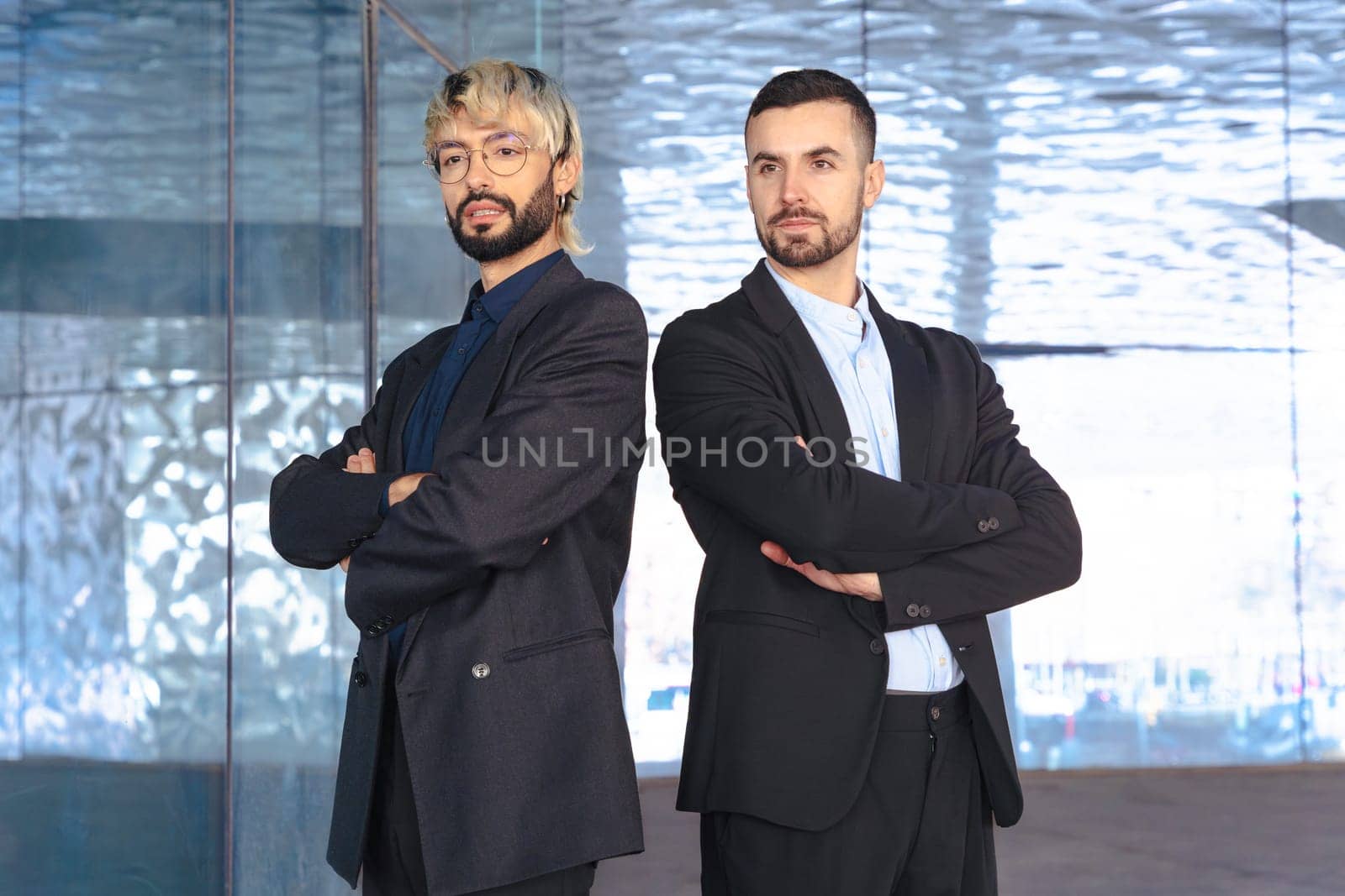 Two confident male professionals in suits standing with crossed arms, serious at the camera inside a modern office. Corporate success, teamwork, and leadership in business environment.
