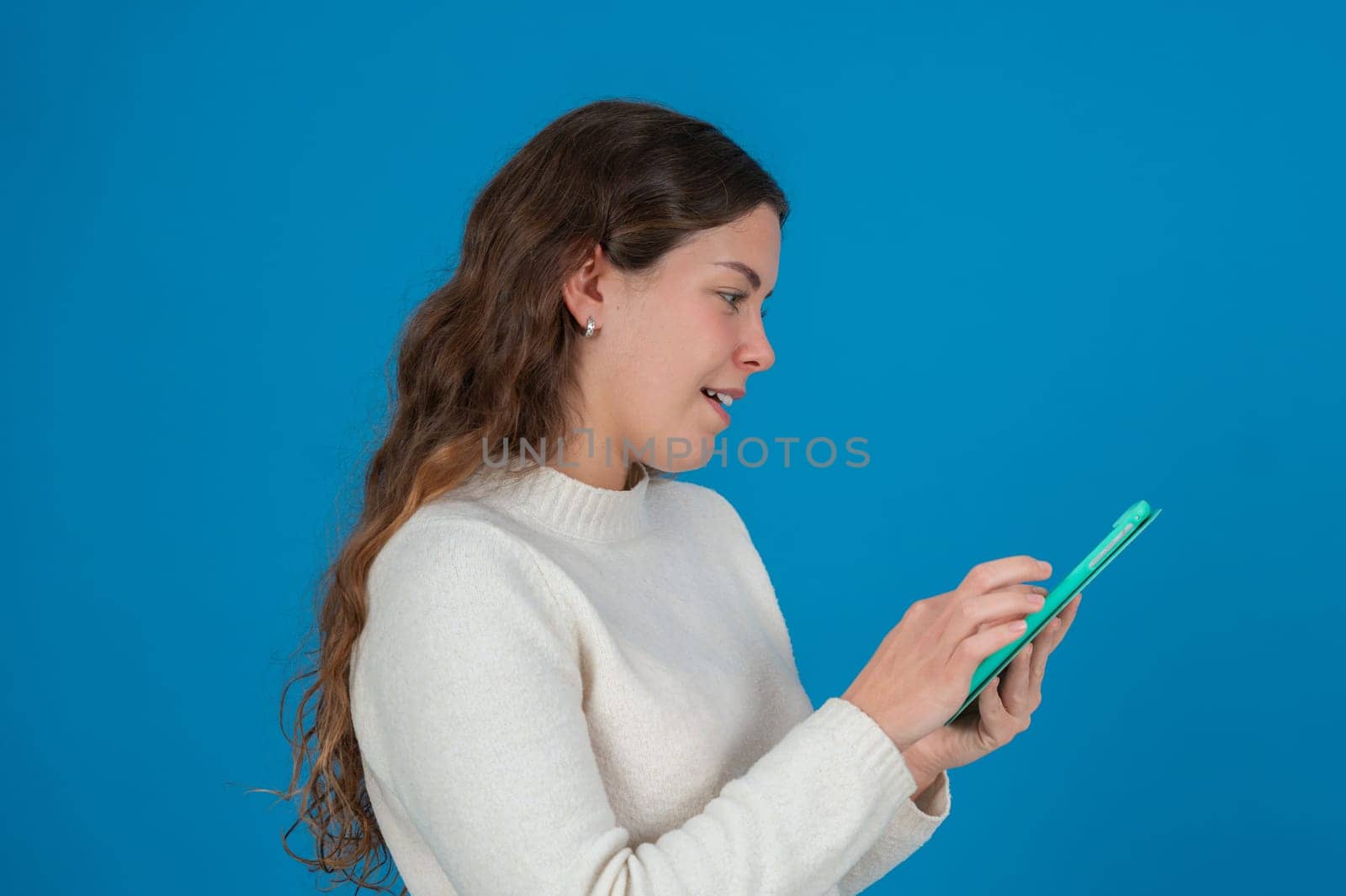 Side view of a smiling young woman in white sweater using a green tablet against a solid blue background with copy space, symbolizing digital communication, browsing, and technology interaction.