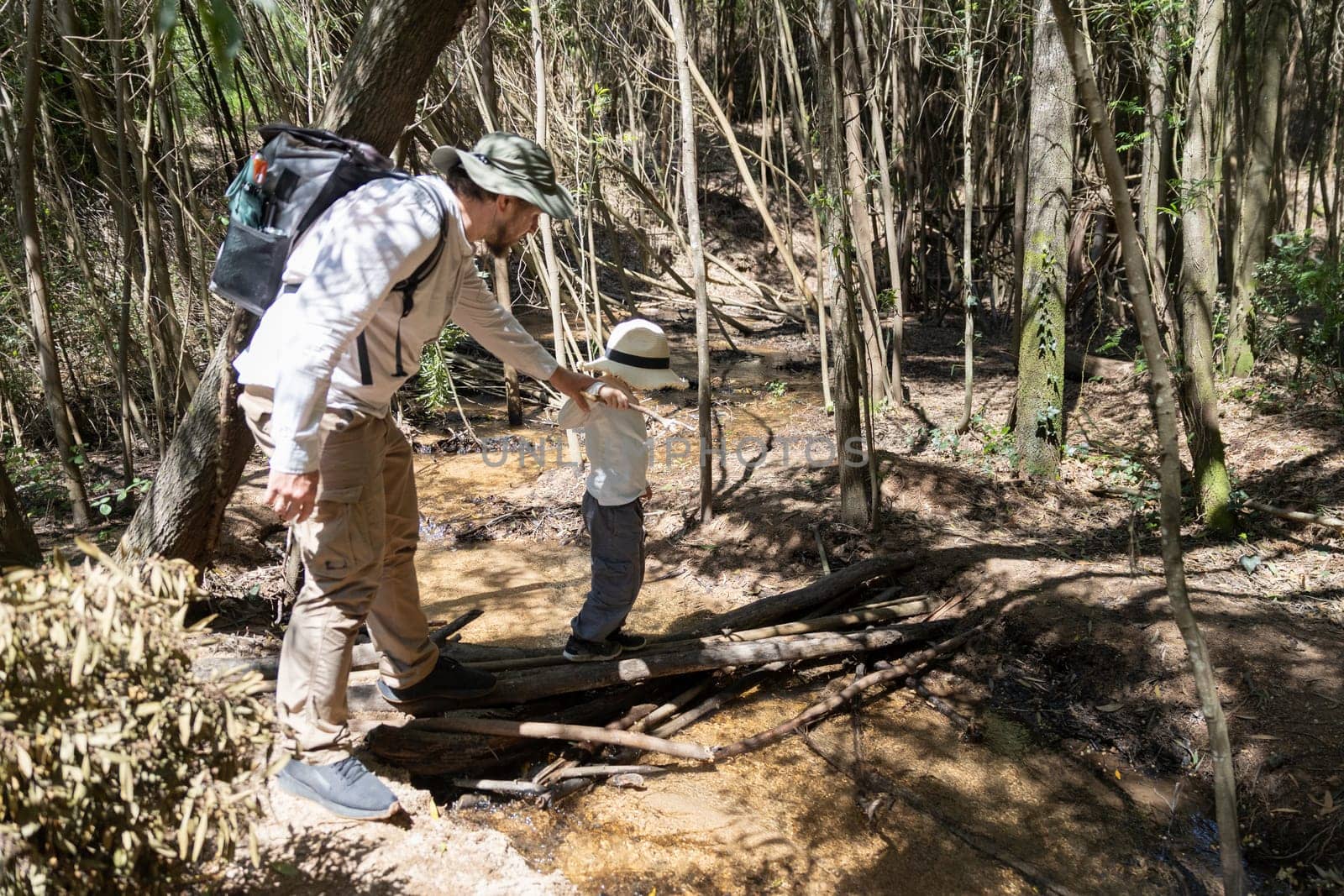 Father helping his son cross a small creek using a makeshift bridge made of branches, during a hike in a sunny forest