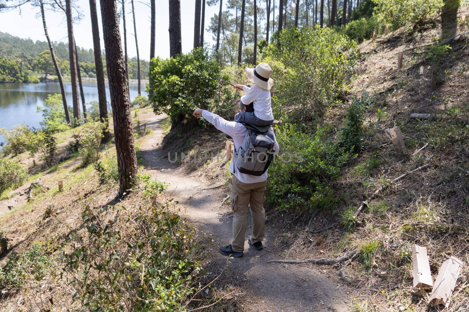 Father carrying daughter on shoulders points across lake while hiking on forest trail