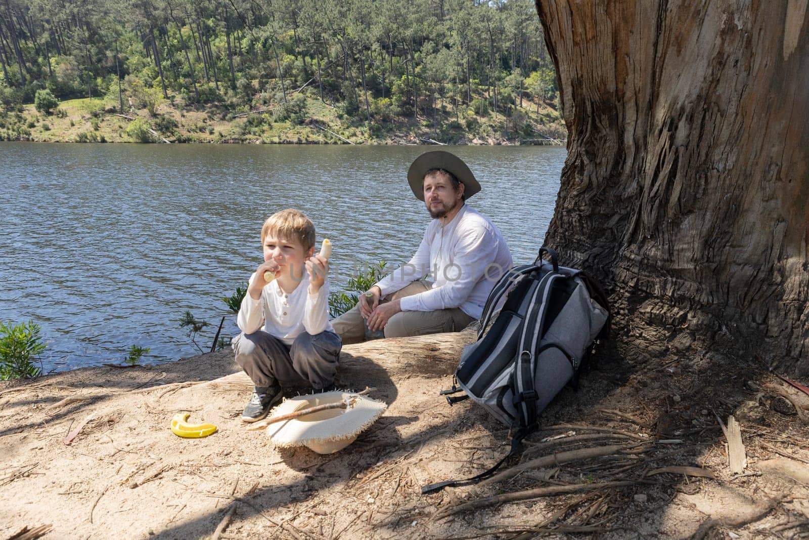 Father and son enjoying a relaxing moment by the lake, eating bananas and resting under a tree