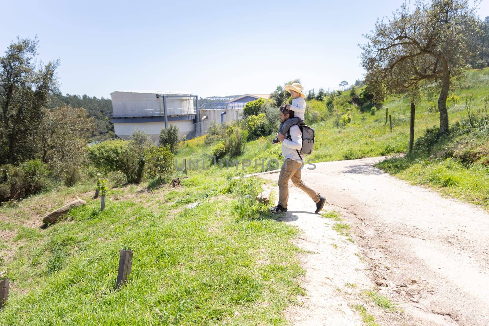 Father carrying daughter on shoulders while walking on country road near water treatment plant