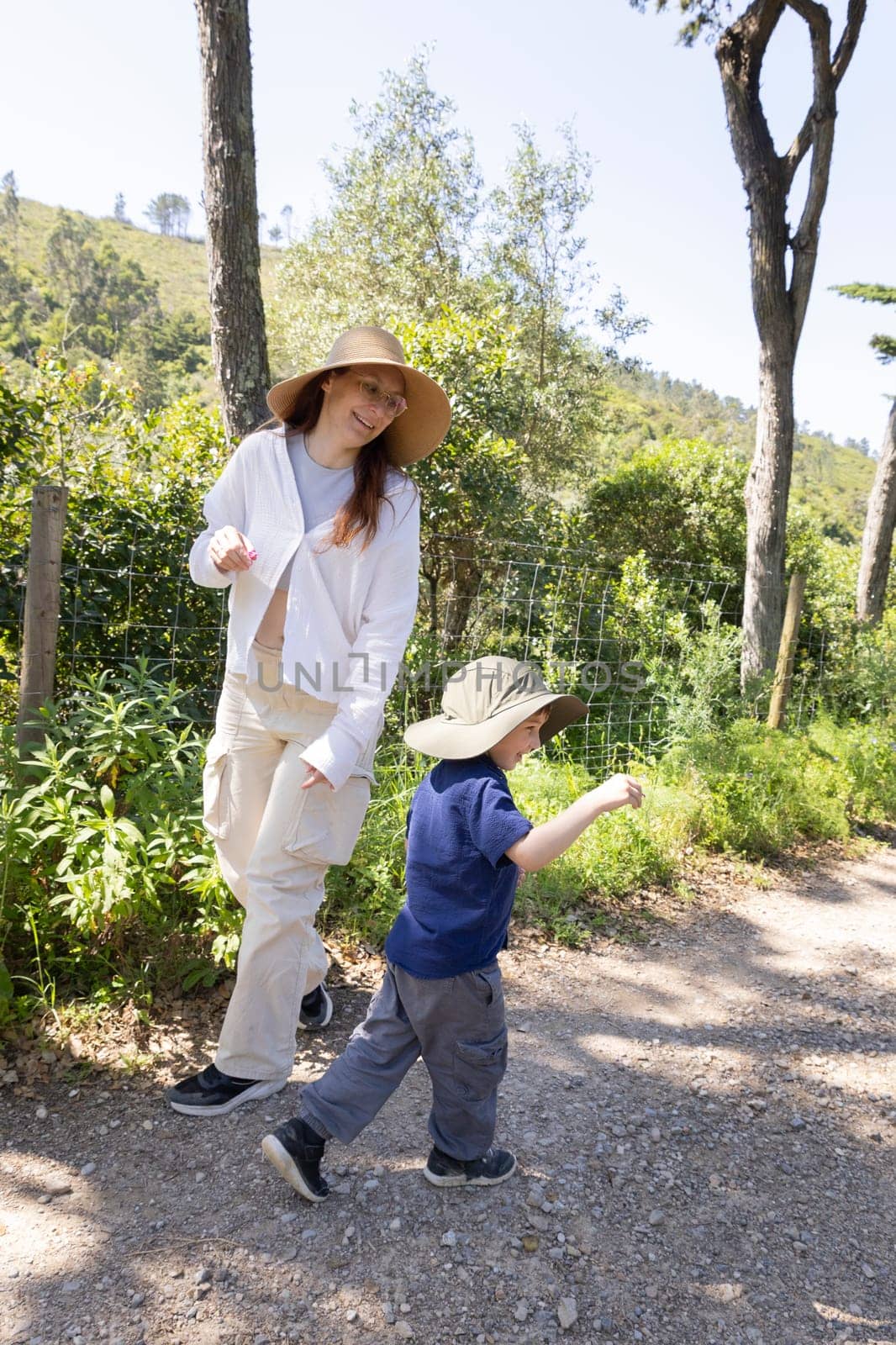 Mother and son wearing hats are walking along a gravel path next to a wire fence, enjoying a sunny day surrounded by nature