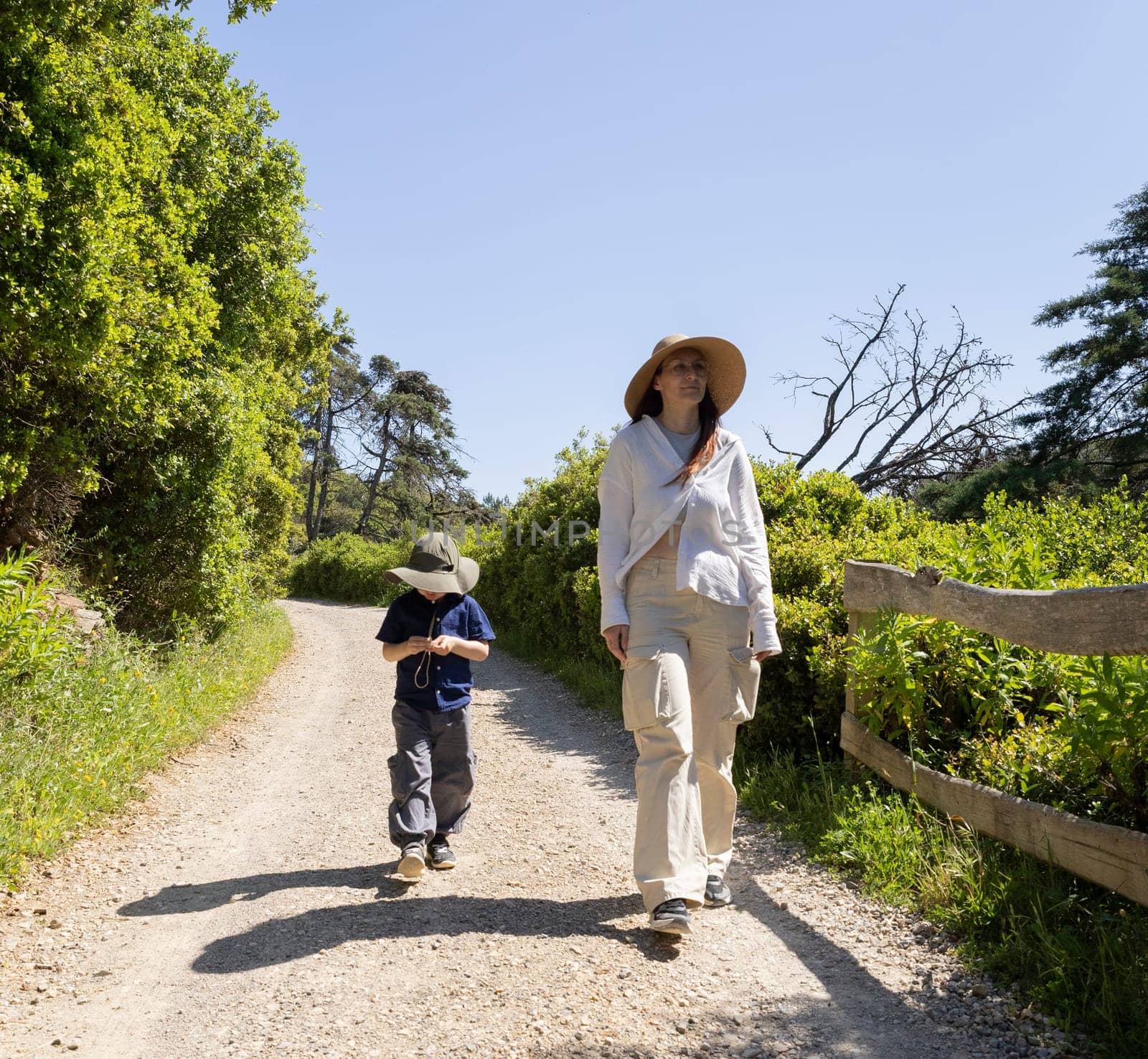 Mother and son wearing hats are walking on a gravel road in nature reserve on a sunny summer day