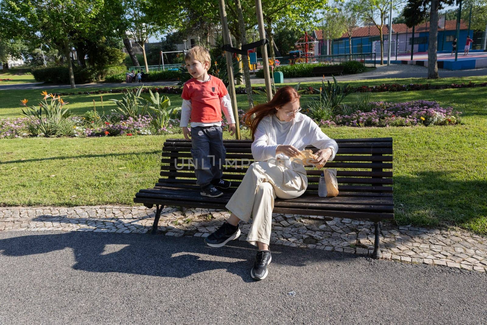 Mother opening a bag of snacks while her son stands on a bench in a park during a sunny spring day