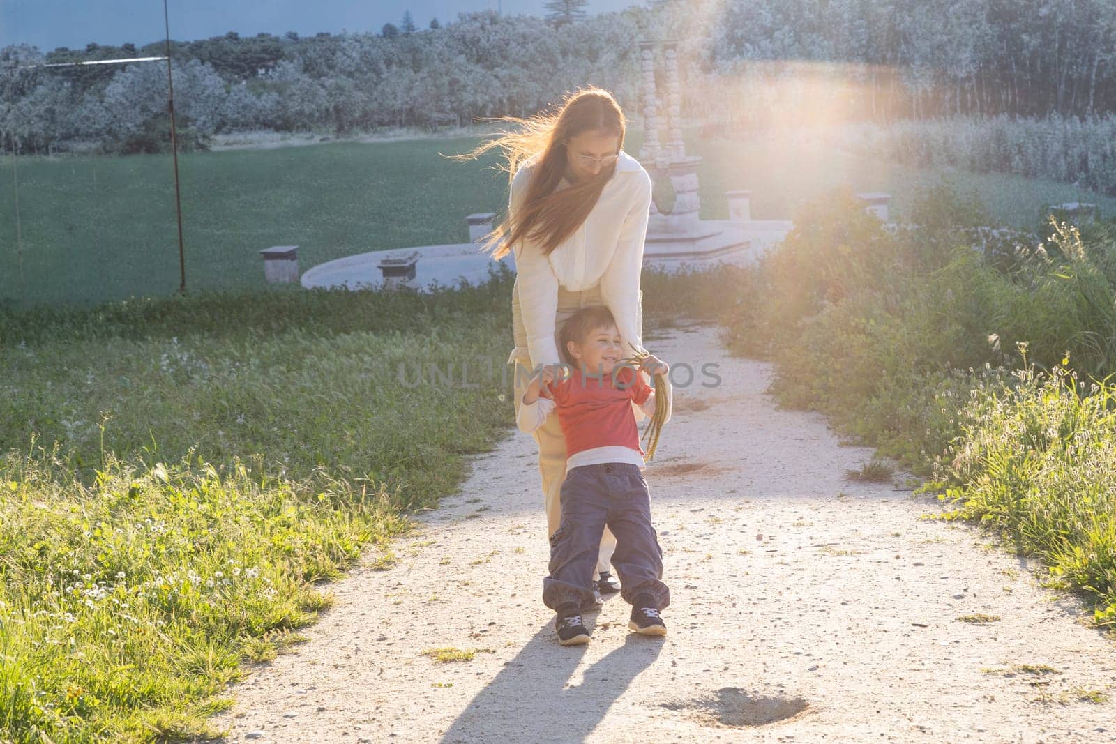 Happy mother playing with her son, swinging him in a beautiful park during a golden sunset