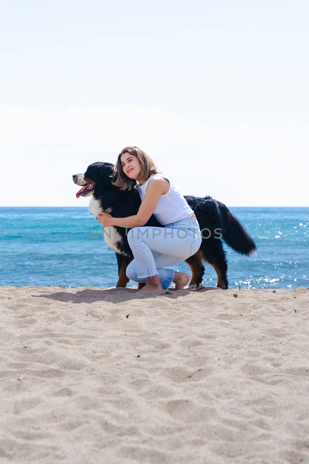 Portrait of a cheerful young woman kneeling beside her dog on a sandy beach. Both look at the camera on a sunny day, enjoying a peaceful moment by the sea with joy, friendship, and nature. Vertical