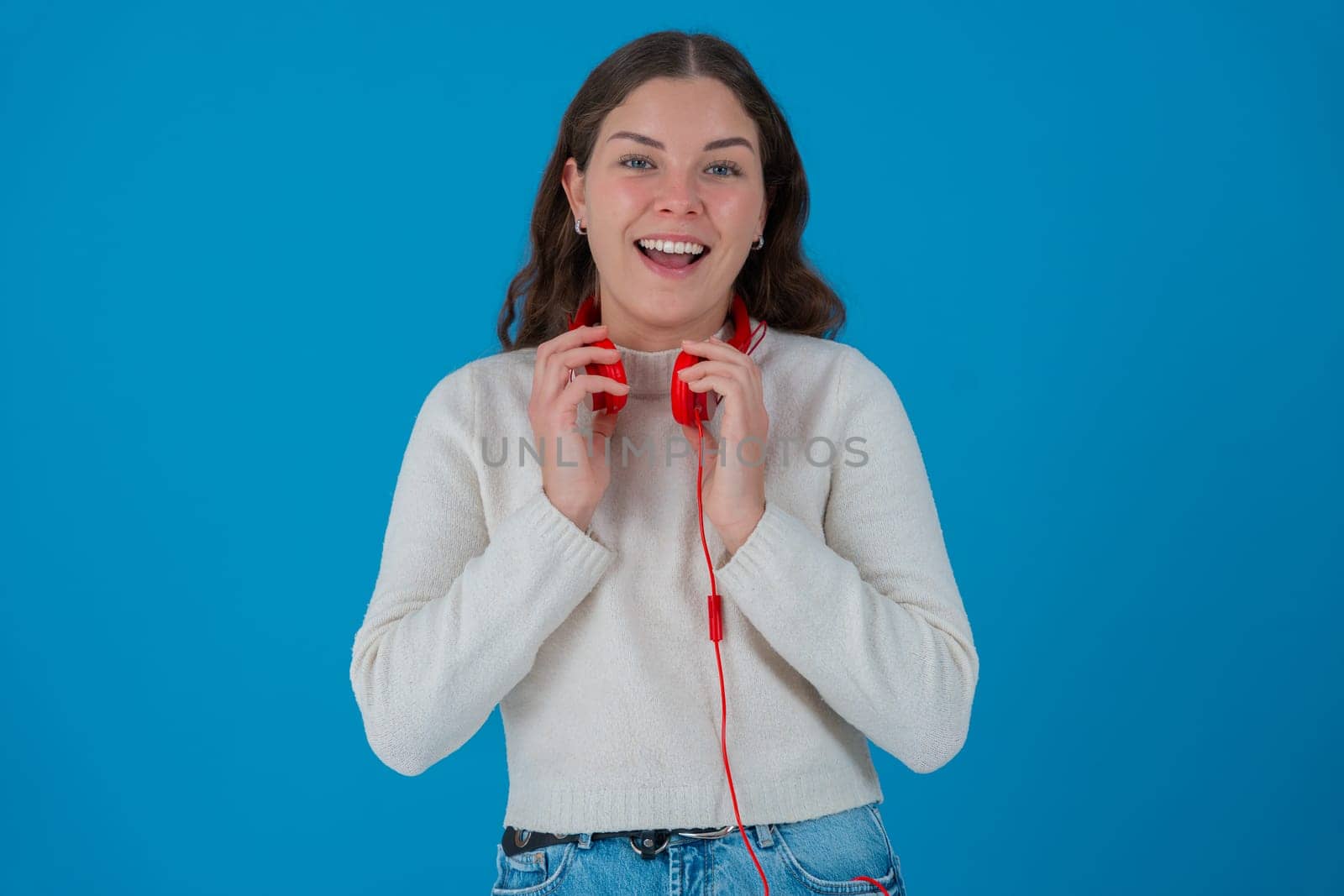 Individuality portrait Young woman wearing red headphones around her neck, smiling happily and looking relaxed. She appears joyful and confident. Solid blue background.