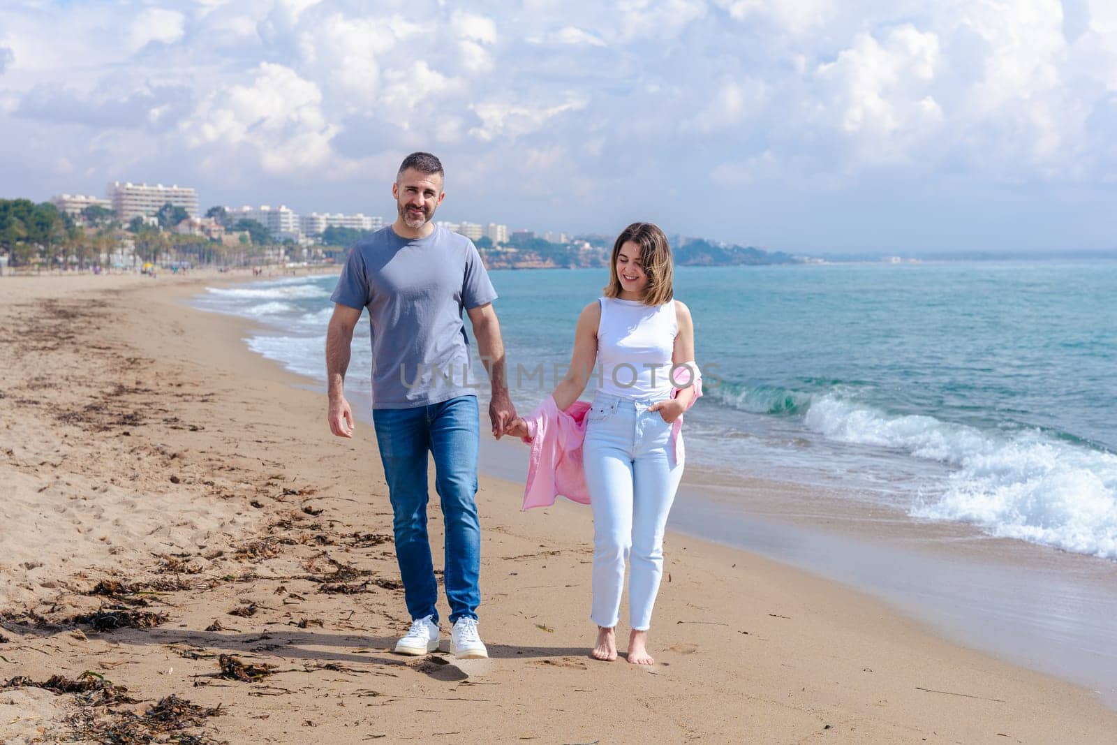 Caucasian young couple walking barefoot on the beach, holding hands and smiling during vacation. Relaxed seaside moment with ocean waves, sand, and a bright sky in the background.copy space
