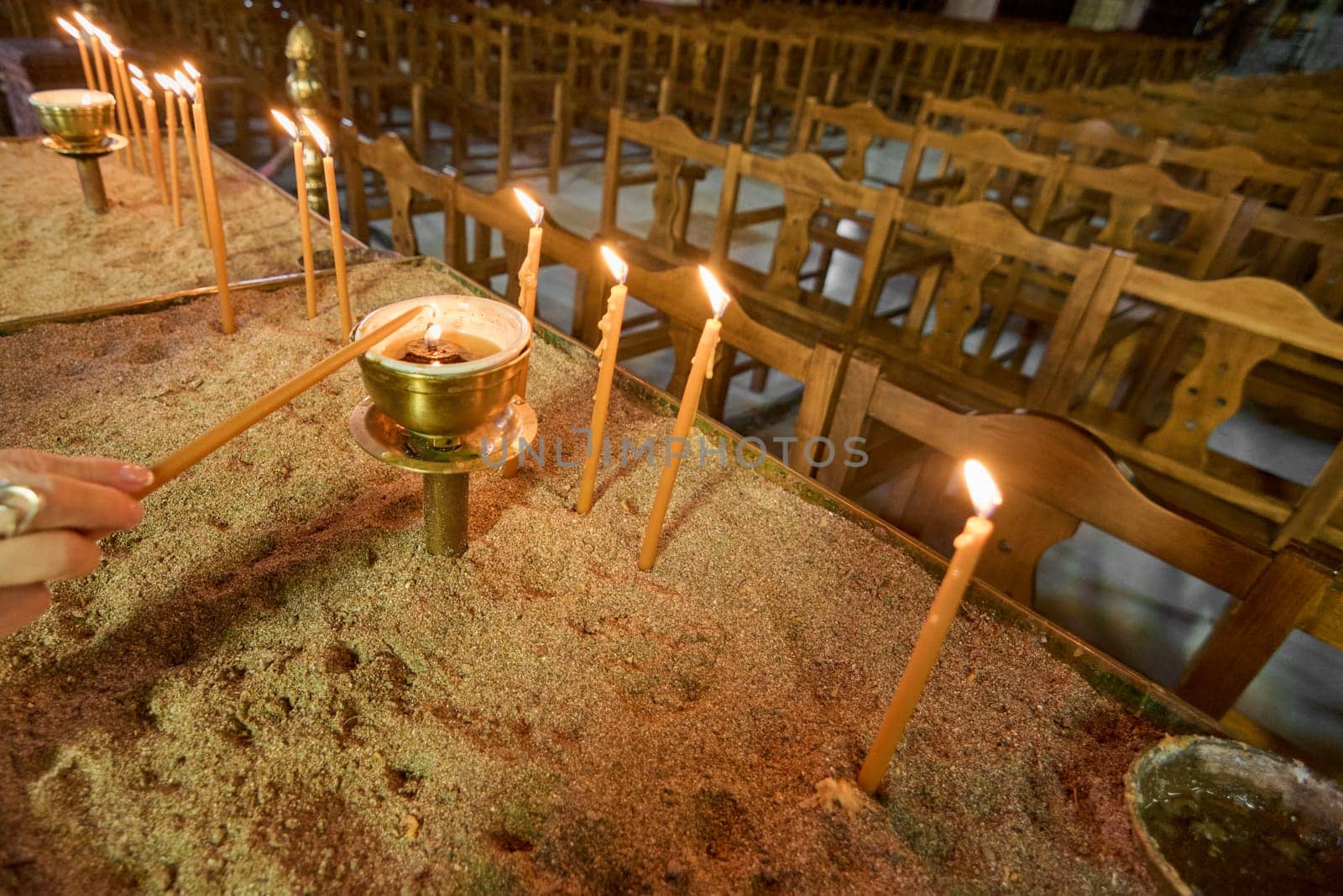 Close-up view of a hand igniting a candle in a sand-filled stand within the serene interior of a Greek Orthodox church. A moment of quiet reverence.