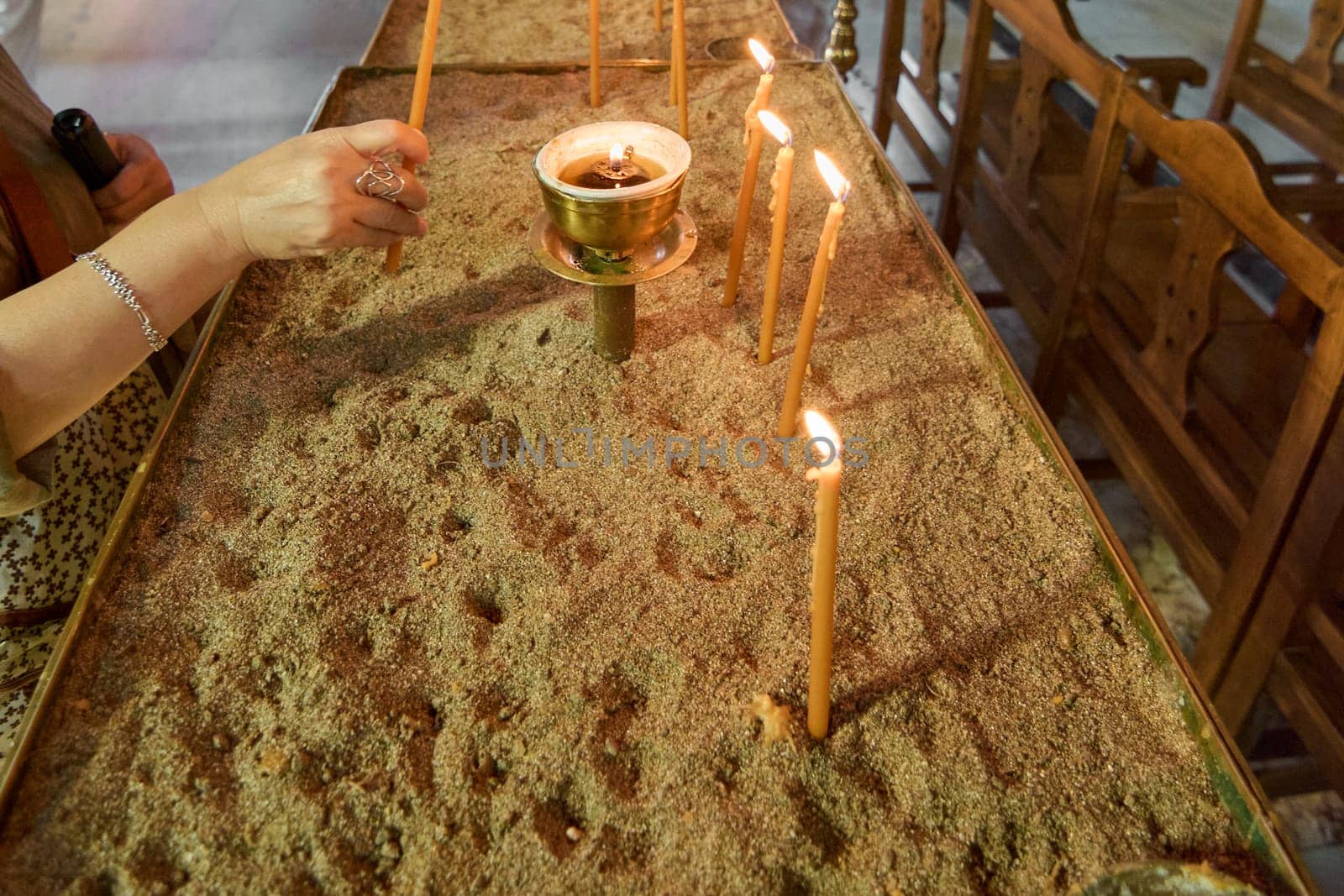A hand lighting a candle in the Church of Annunciation, a symbolic act of prayer and spiritual devotion, with rows of wooden benches in the background.