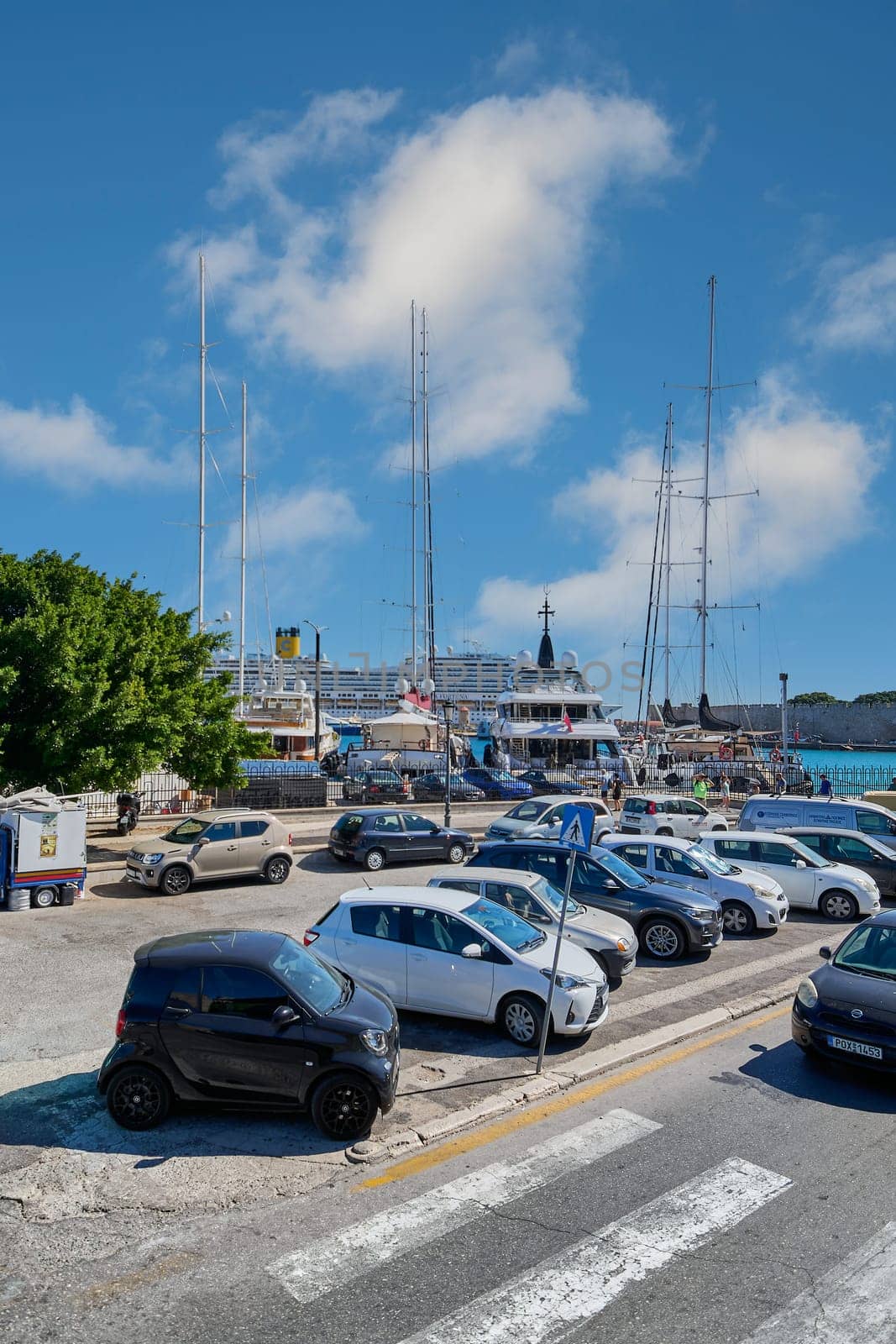Rodas.Greece - june 05, 2025: View of a street with parked vehicles and the vibrant Rhodes port. The intersection of urban life and maritime activity.