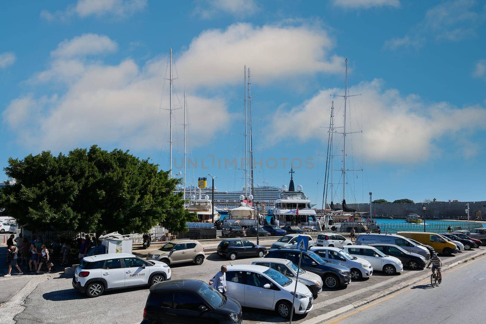 Rodas.Greece - june 05, 2025: A parking lot with various cars and a motorcycle, featuring Mandraki port and vessels in the background. Everyday city scene.