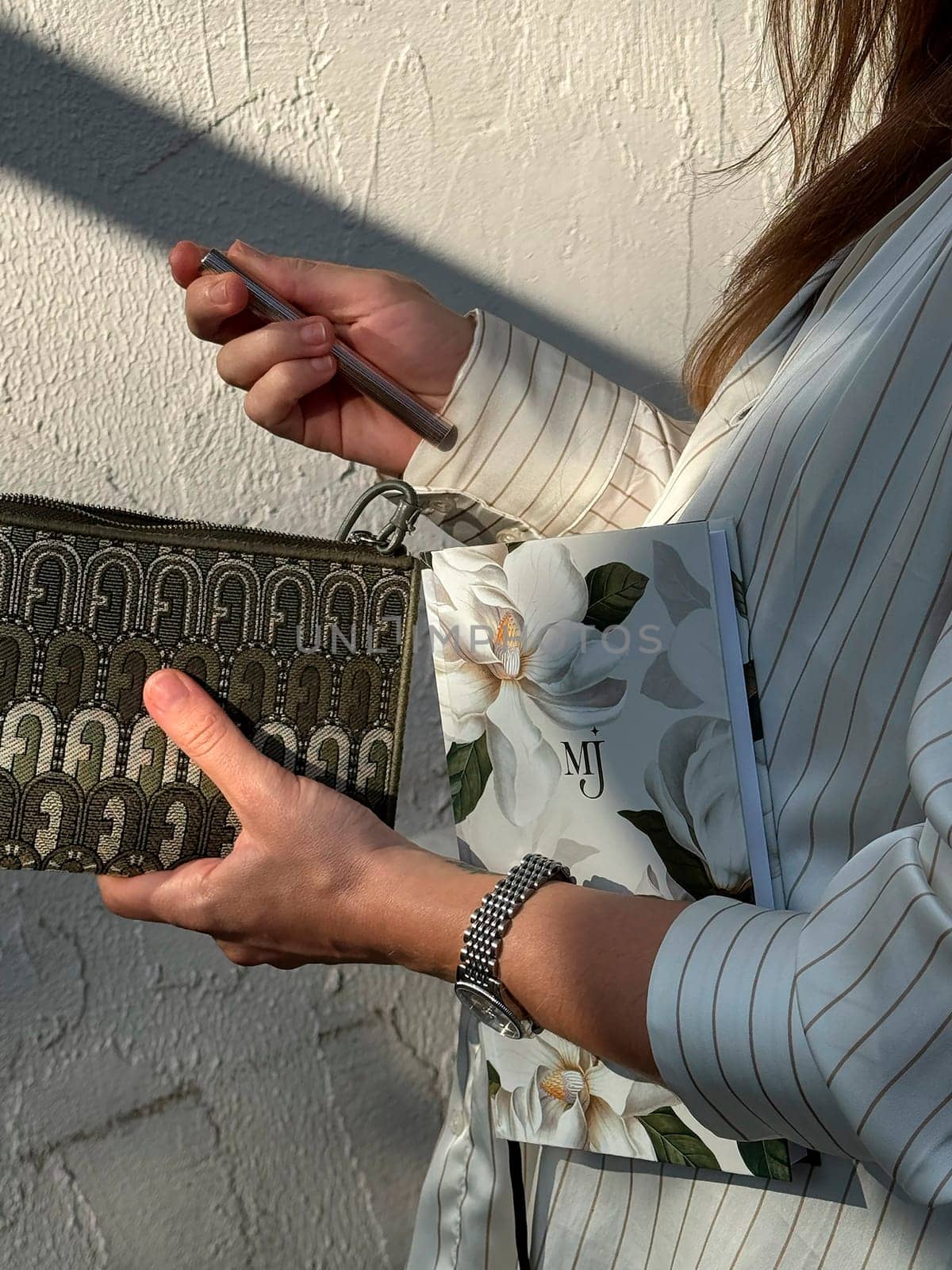 A woman dressed in a striped shirt stands outdoors, clutching a decorative wallet and a floral notepad in natural light.