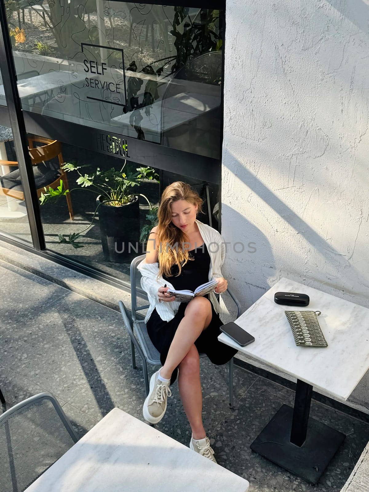 Woman with long hair reads a book at a stylish cafe table surrounded by bright indoor plants in the afternoon light.
