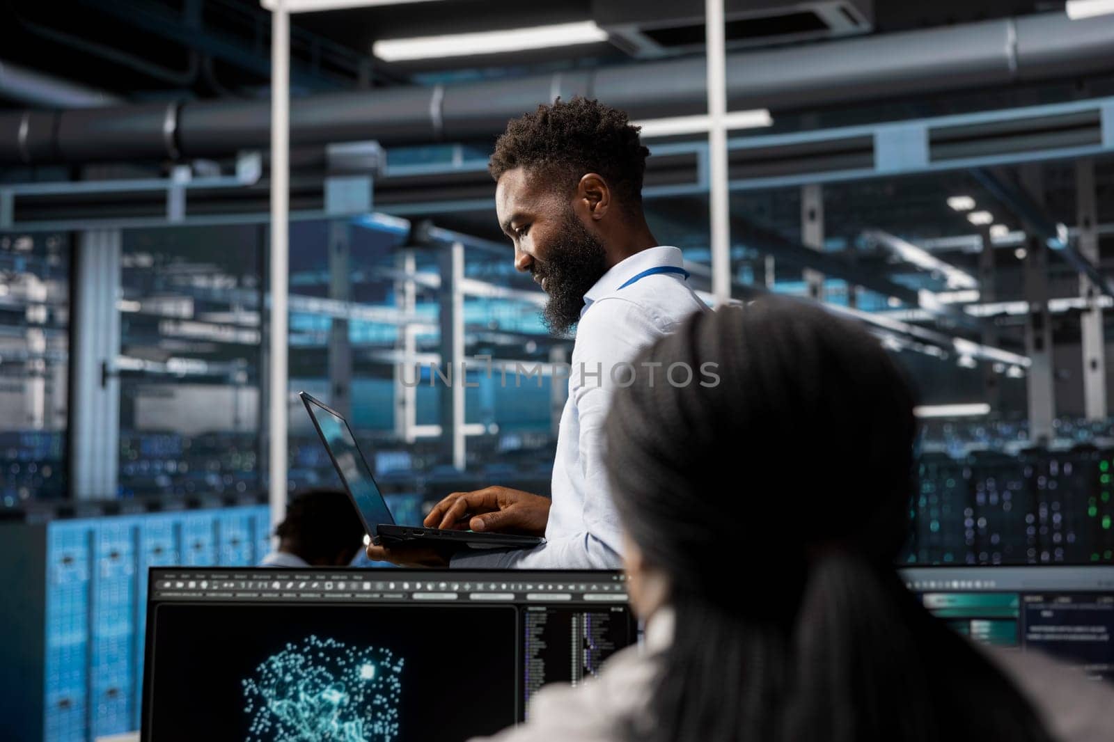 Data center system administrator uses AI visualization tech to oversee automated repetitive tasks. African american man in server farm modifying infrastructure rigs based on data analytics graphs