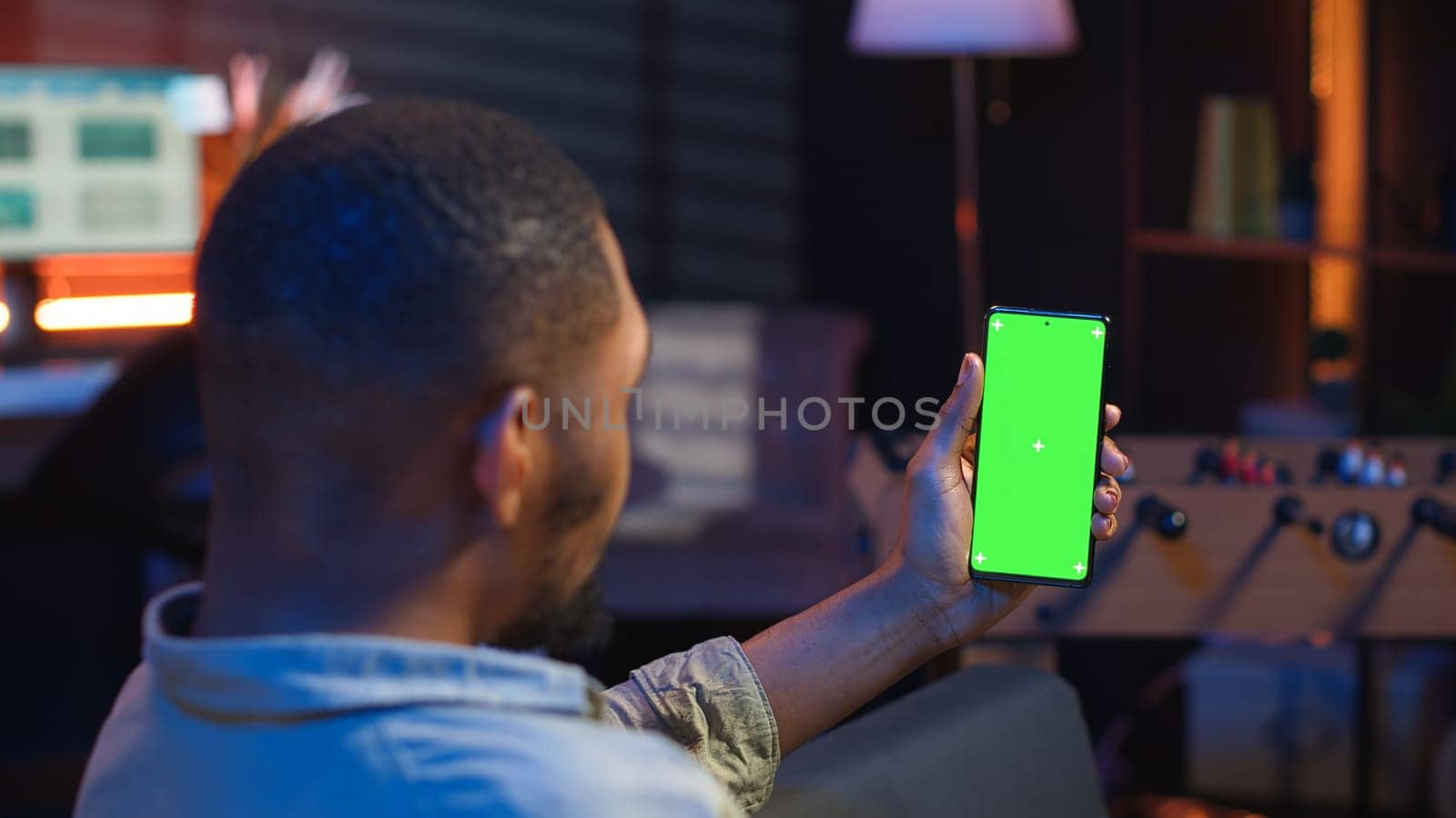 African american young adult laying on the sofa with mockup screen, feeling happy and relaxed with flexible work schedule at home. Freelancer uses phone relaxing in the living room. Camera B.