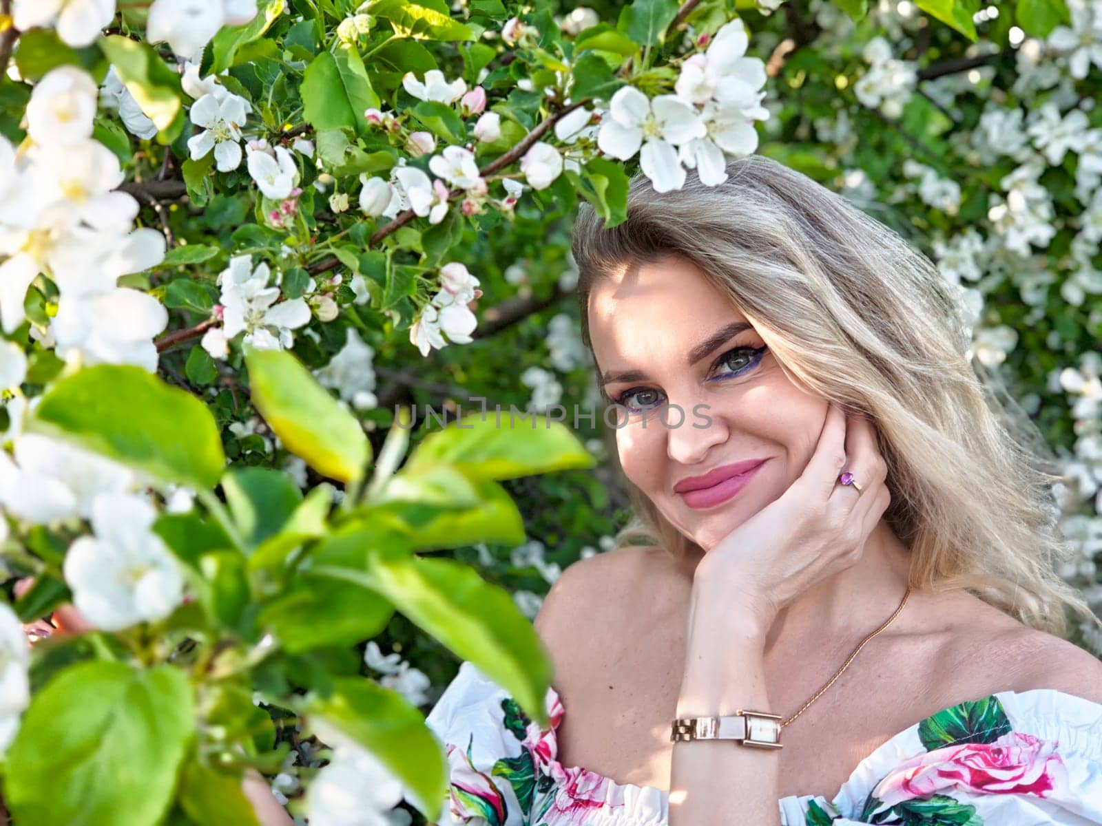 A mature blonde woman poses among blooming flowers, capturing a joyful moment for her lifestyle blog in a sunny garden.