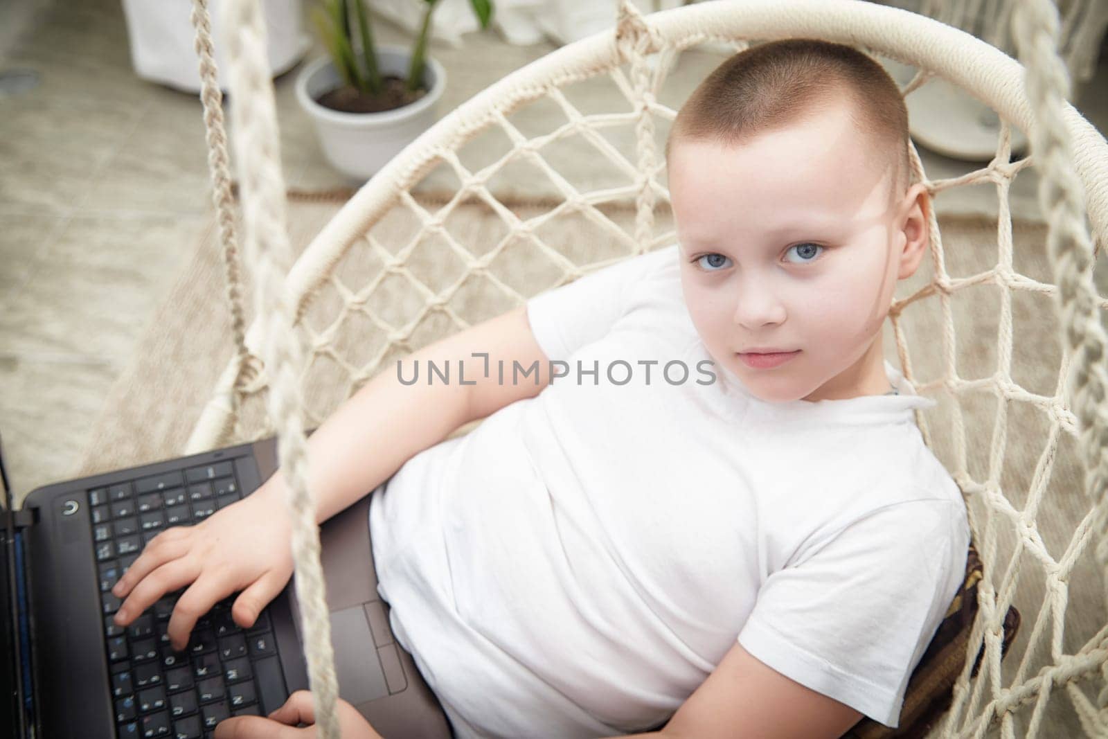 A young boy in a white shirt comfortably lounges in a hanging chair, focused on his laptop in a bright indoor setting.
