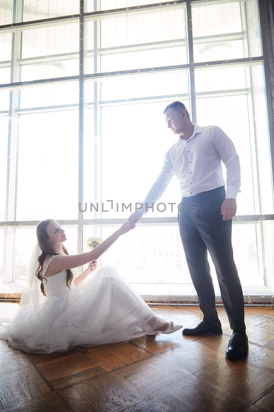 Groom and bride share a tender moment while seated on the floor, bathed in natural light from large windows during their wedding.