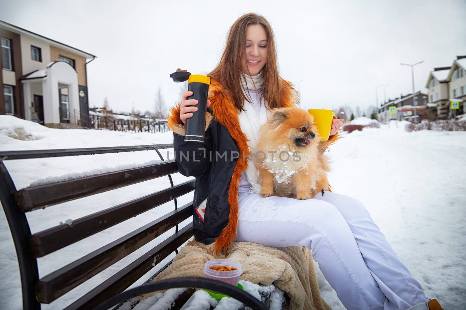 A woman sits on a bench, holding a drink and snacks while her fluffy Pomeranian rests beside her in the snow.