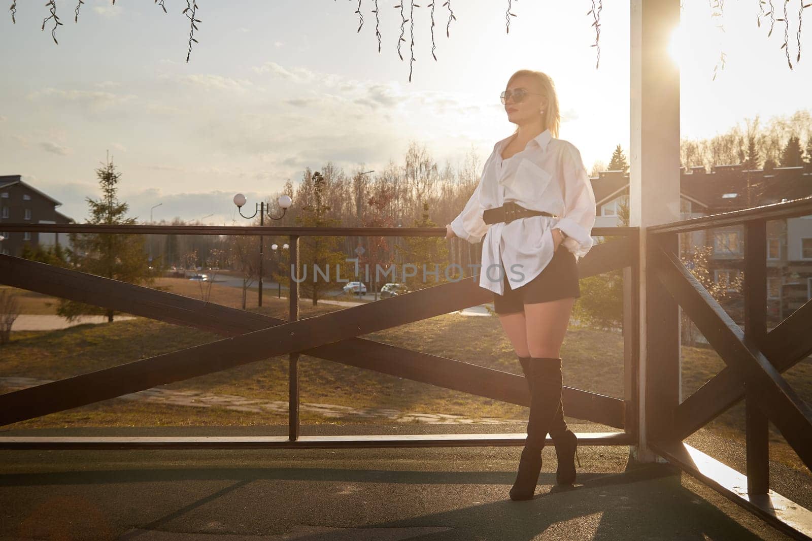 Beautiful girl with red hair in white shirt in open wooden pavillion in village or small town. Young slender woman and sky with clouds on background on autumn, spring or summer day