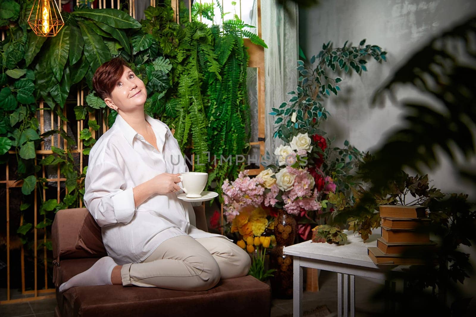 Middle-aged woman sits comfortably in a cozy room, sipping tea and surrounded by lush plants and decor.