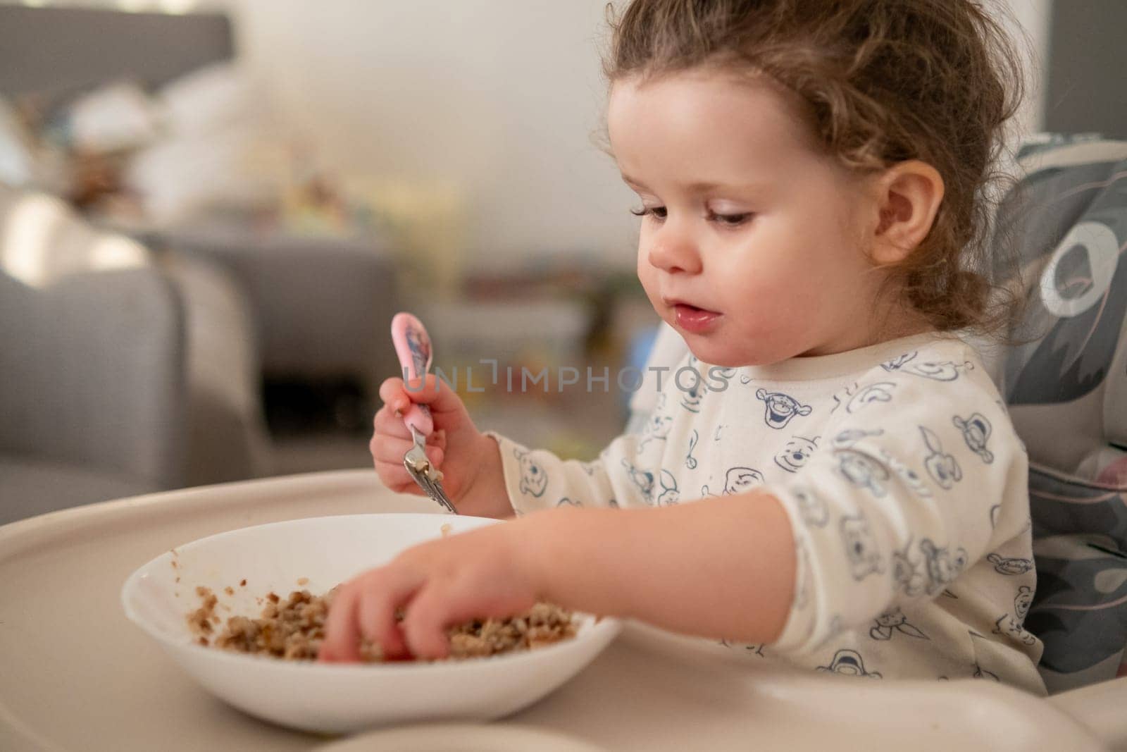 Little girl with curly hair focused on eating porridge in the kitchen. Messy and sweet moment of toddler mealtime.