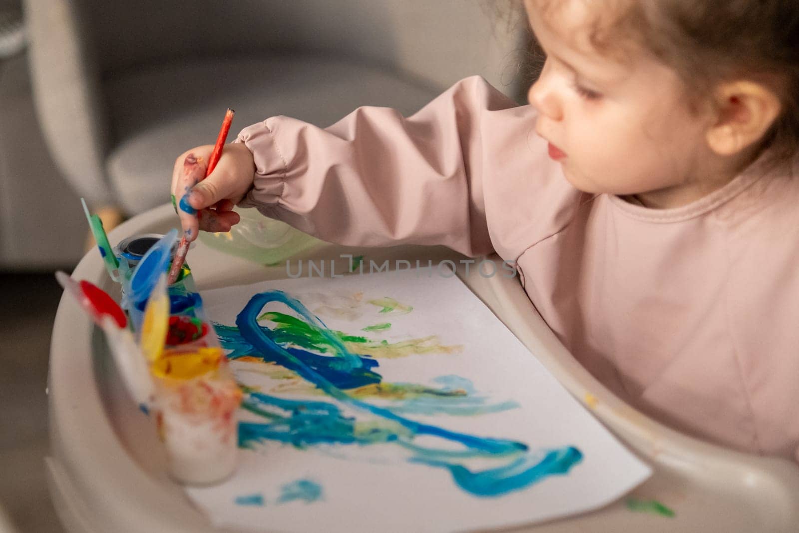 Child drawing with gouache while sitting on a colorful plastic chair. Early childhood creativity and art education at home.