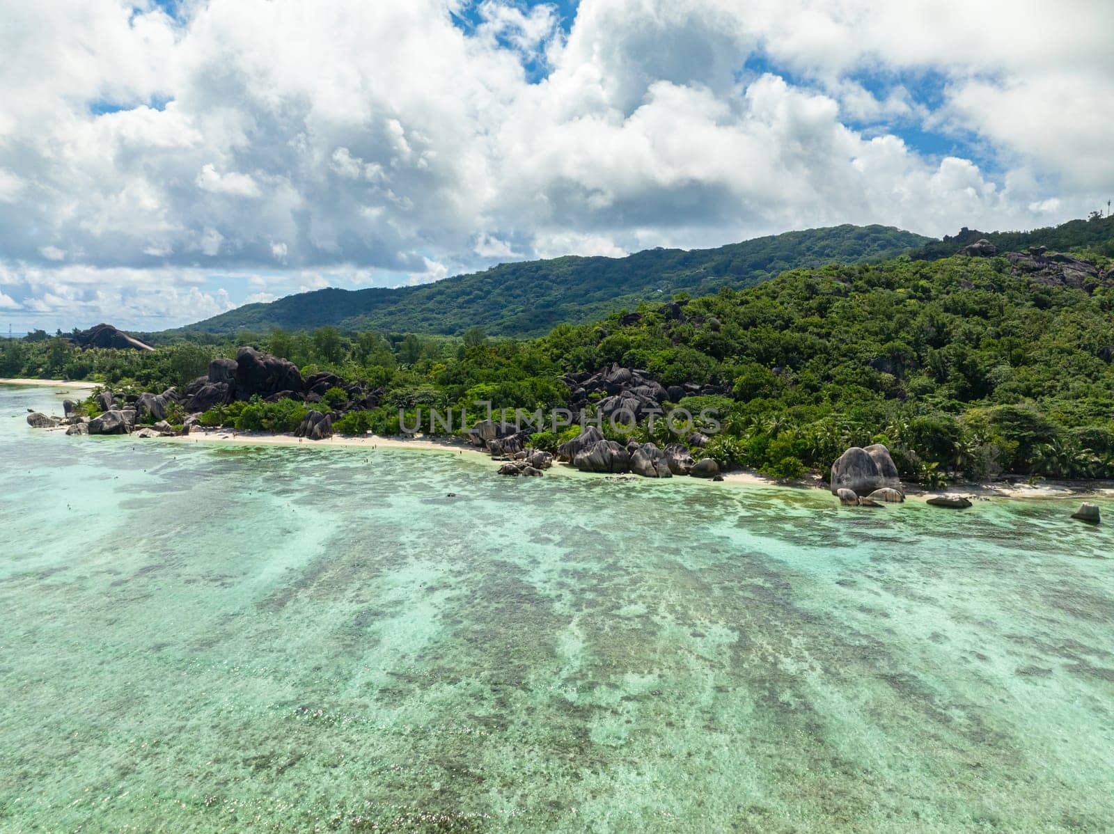 Aerial shot of lush green hills meeting the coast with cloudy skies overhead. La Digue, Seychelles.