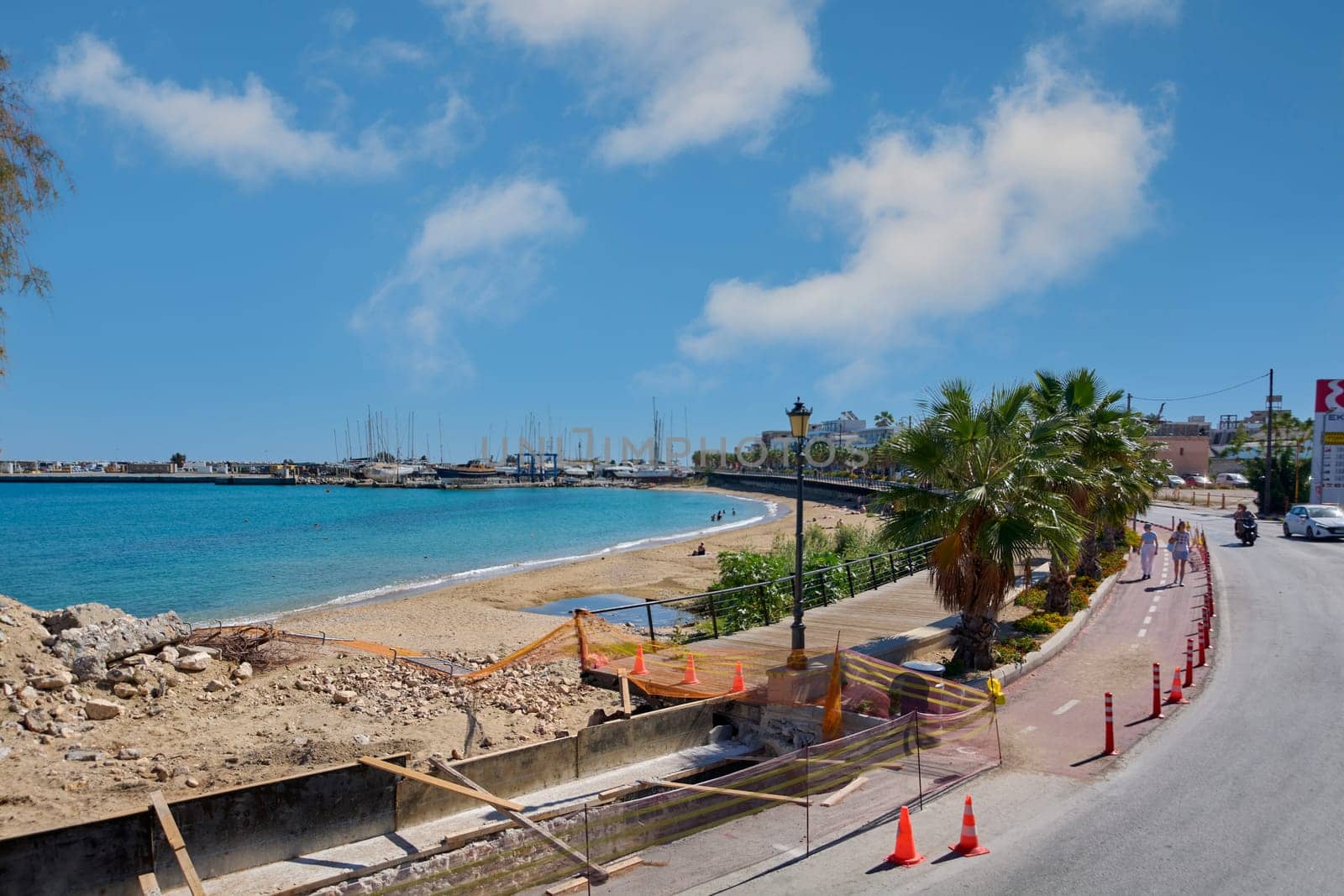 Rodas.Greece - june 06, 2025: A view of Rhodes beach with people enjoying the sun, a promenade under construction, and the harbor full of boats in the background. Blue sky with white clouds.