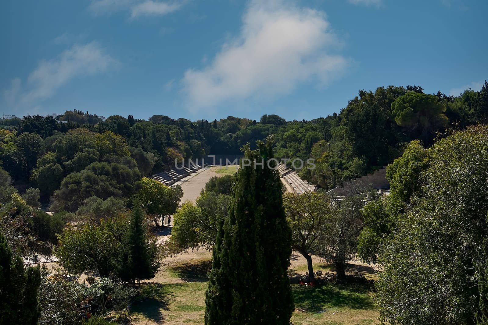 A panoramic view of the ancient stadium at the Acropolis of Rhodes, Greece, showcasing the ruins and abundant greenery under a clear blue sky.