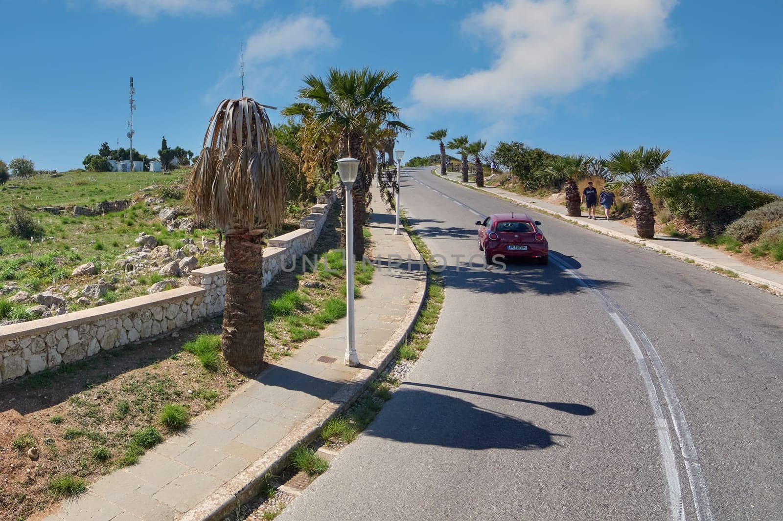 Rhodes. Greece - june 06, 2025: A road ascends in Rhodes, Greece, flanked by palm trees and a stone wall, with a red car and a blue sky, reflecting the island's charm.