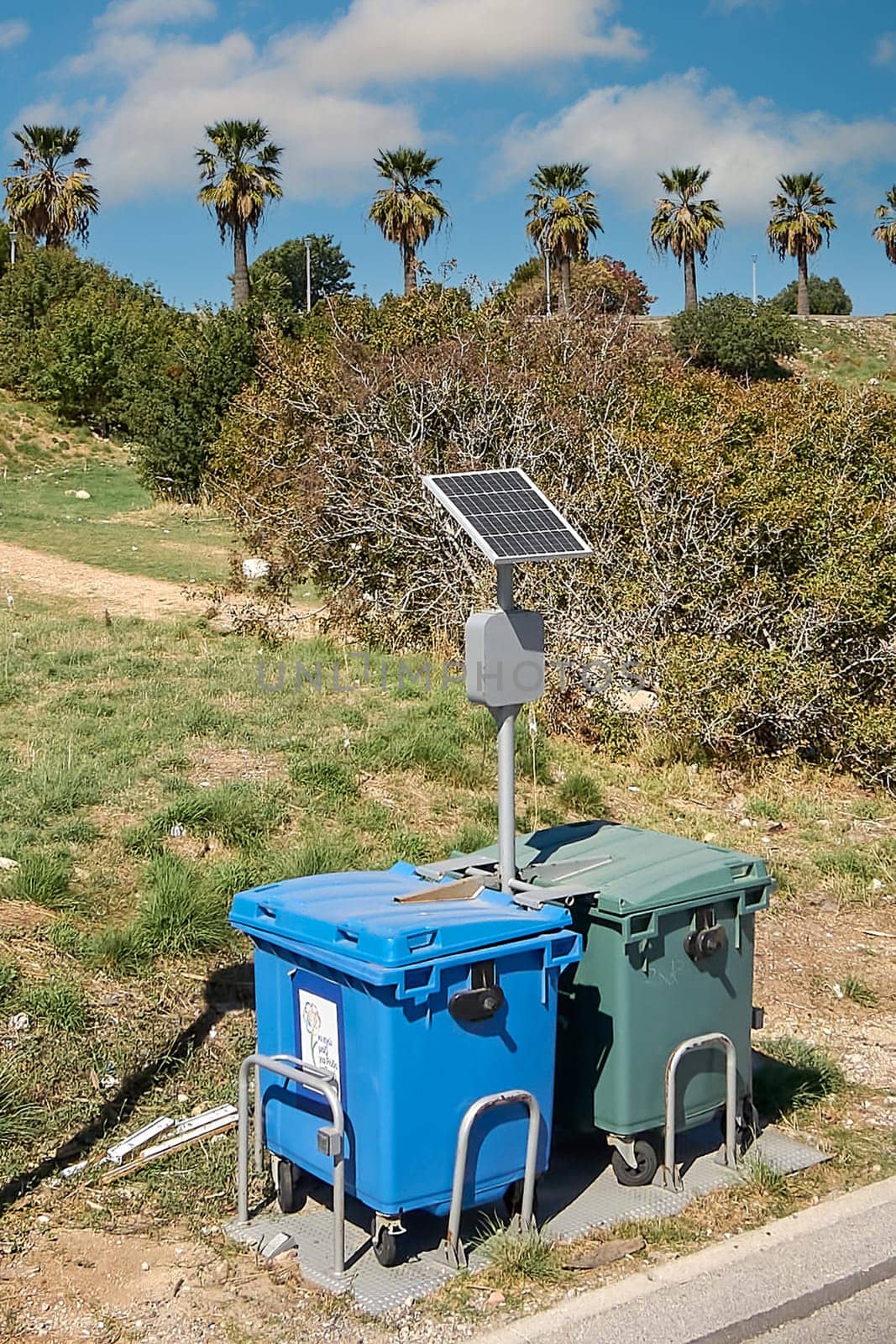 Recycling bins with a solar panel in a natural setting in Rhodes, Greece, with palm trees in the background, symbolizing sustainability and waste management.
