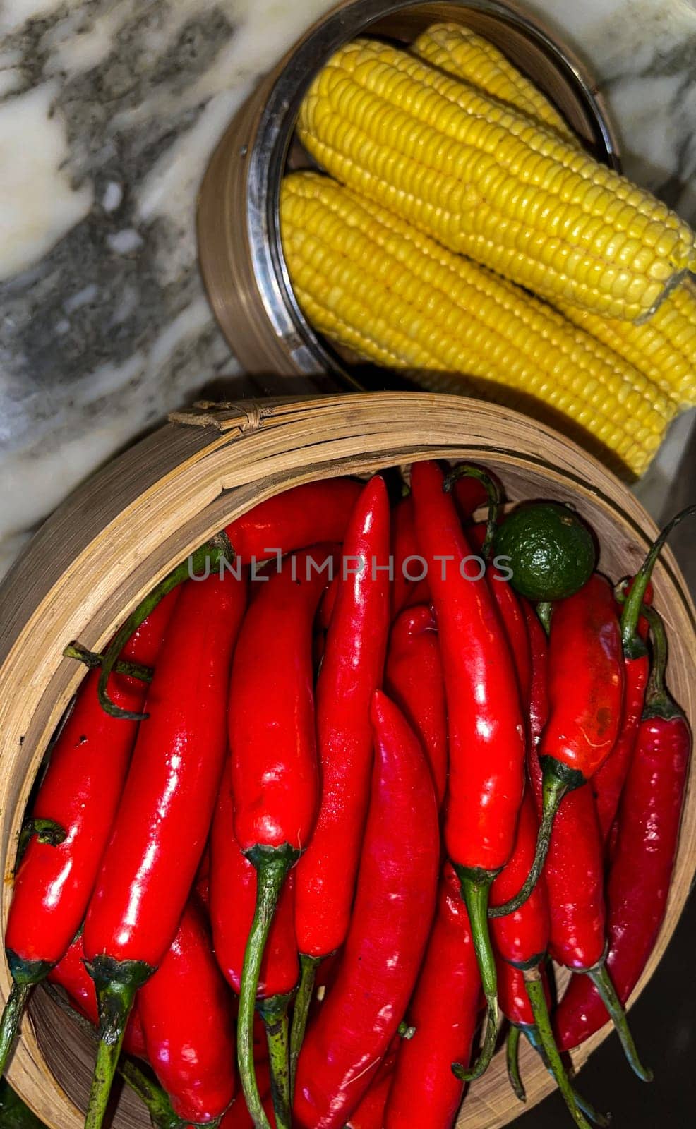 Vibrant red chili peppers with a single green lime resting in a wooden bowl beside yellow corn cobs on a stone surface, enhancing the natural and fresh themes of the composition.