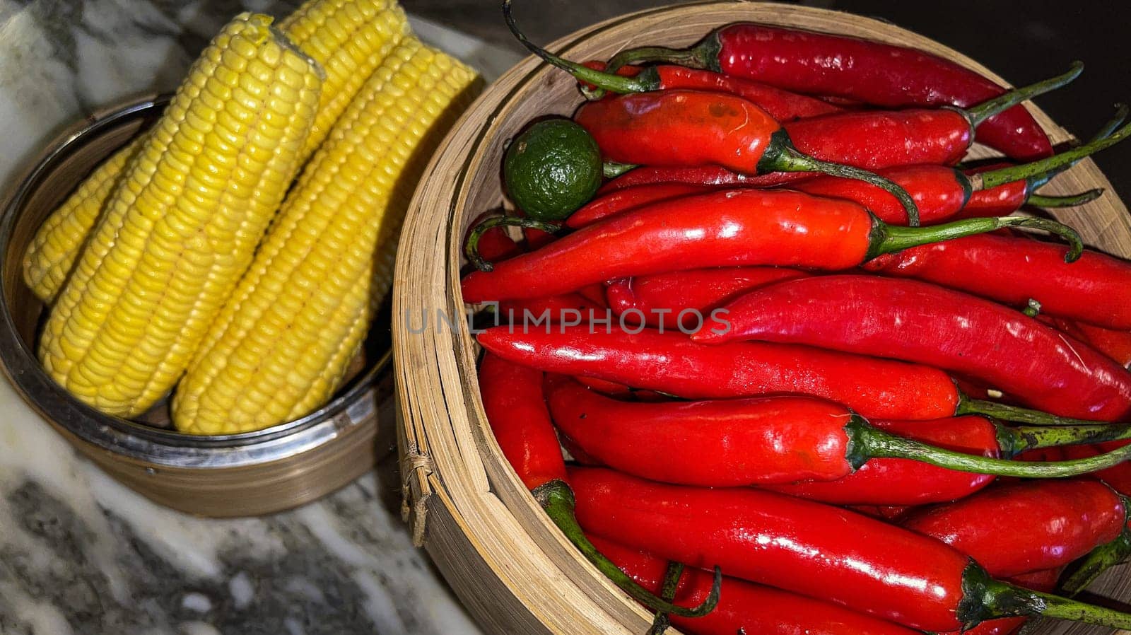 Vibrant red chili peppers with a single green lime resting in a wooden bowl beside yellow corn cobs on a stone surface, enhancing the natural and fresh themes of the composition.