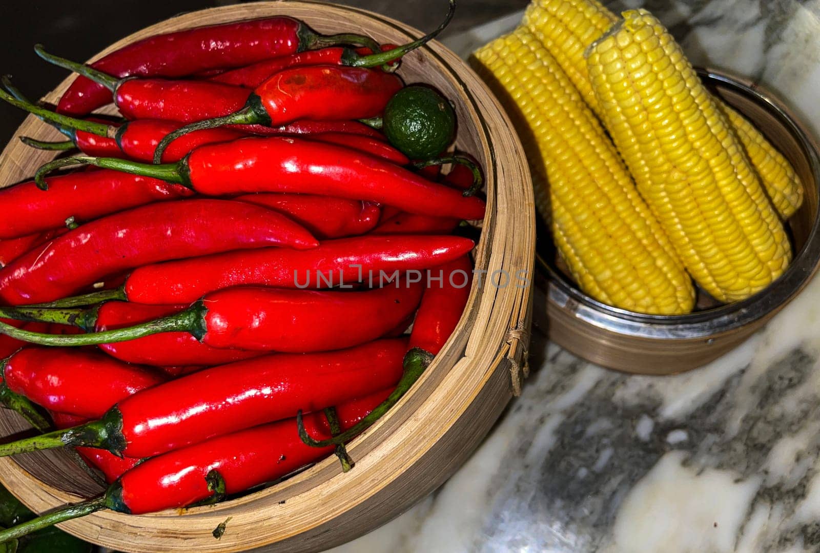 Vibrant red chili peppers with a single green lime resting in a wooden bowl beside yellow corn cobs on a stone surface, enhancing the natural and fresh themes of the composition.