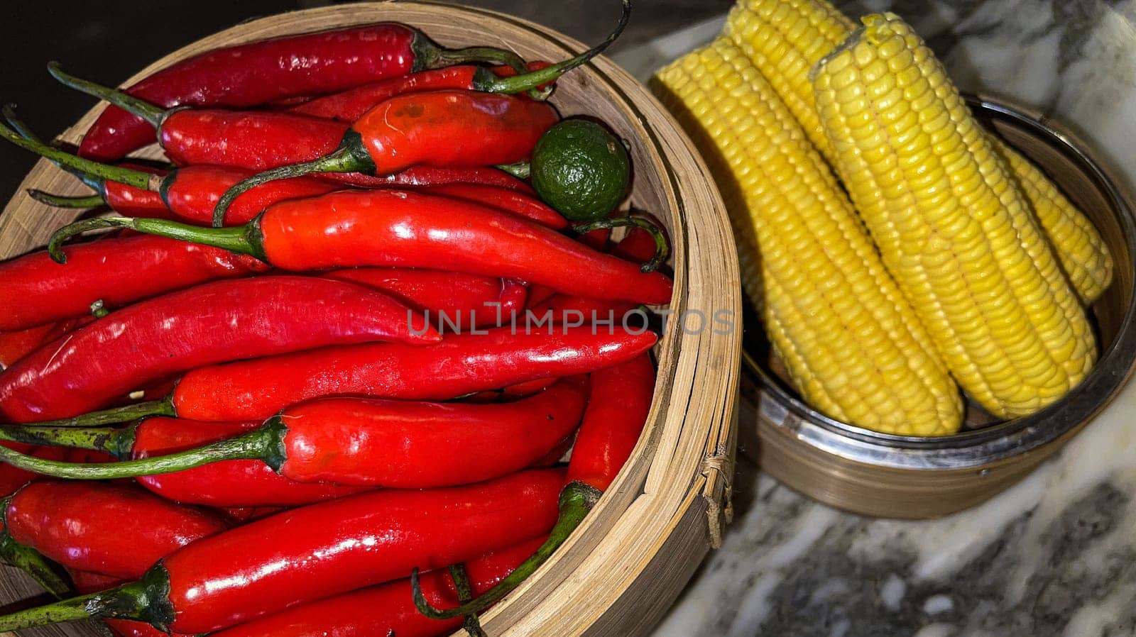Vibrant red chili peppers with a single green lime resting in a wooden bowl beside yellow corn cobs on a stone surface, enhancing the natural and fresh themes of the composition.
