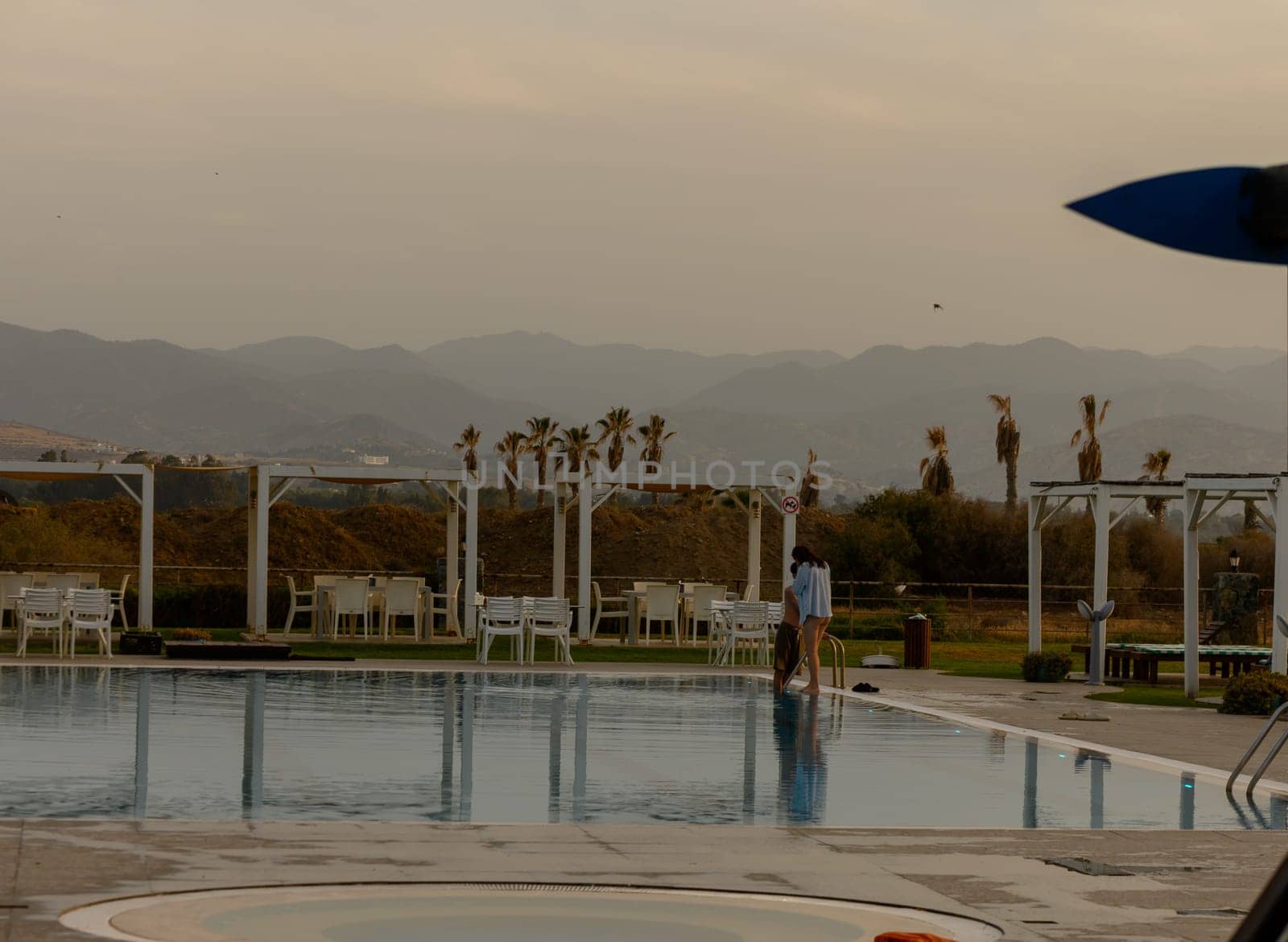 A young man and woman share a warm embrace beside a tranquil swimming pool in Cyprus, surrounded by serene holiday vibes.