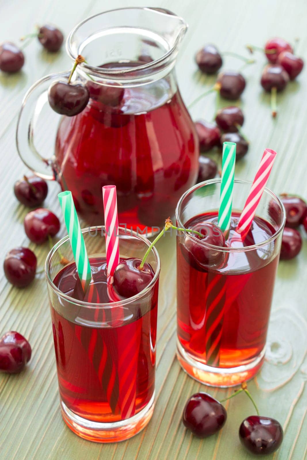 Two glasses of cherry juice with straws in the foreground and in the background a jar full of cherry juice. 2 cherry juices. Vertical. Close-up. Daylight.