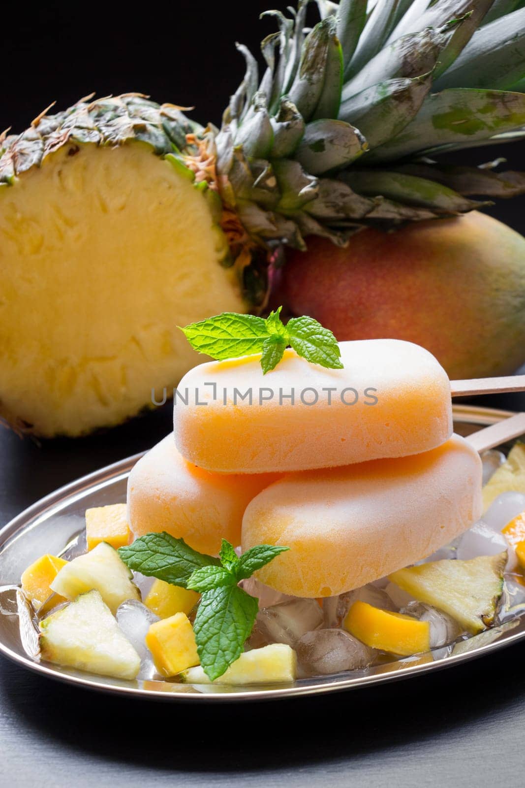 Exotic sorbet ice cream popsicles on silver plate with ice cubes chopped pineapple mango pieces, mint near half of pineapple, mango on a black background. Pineapple mango sorbet ice cream popsicles.