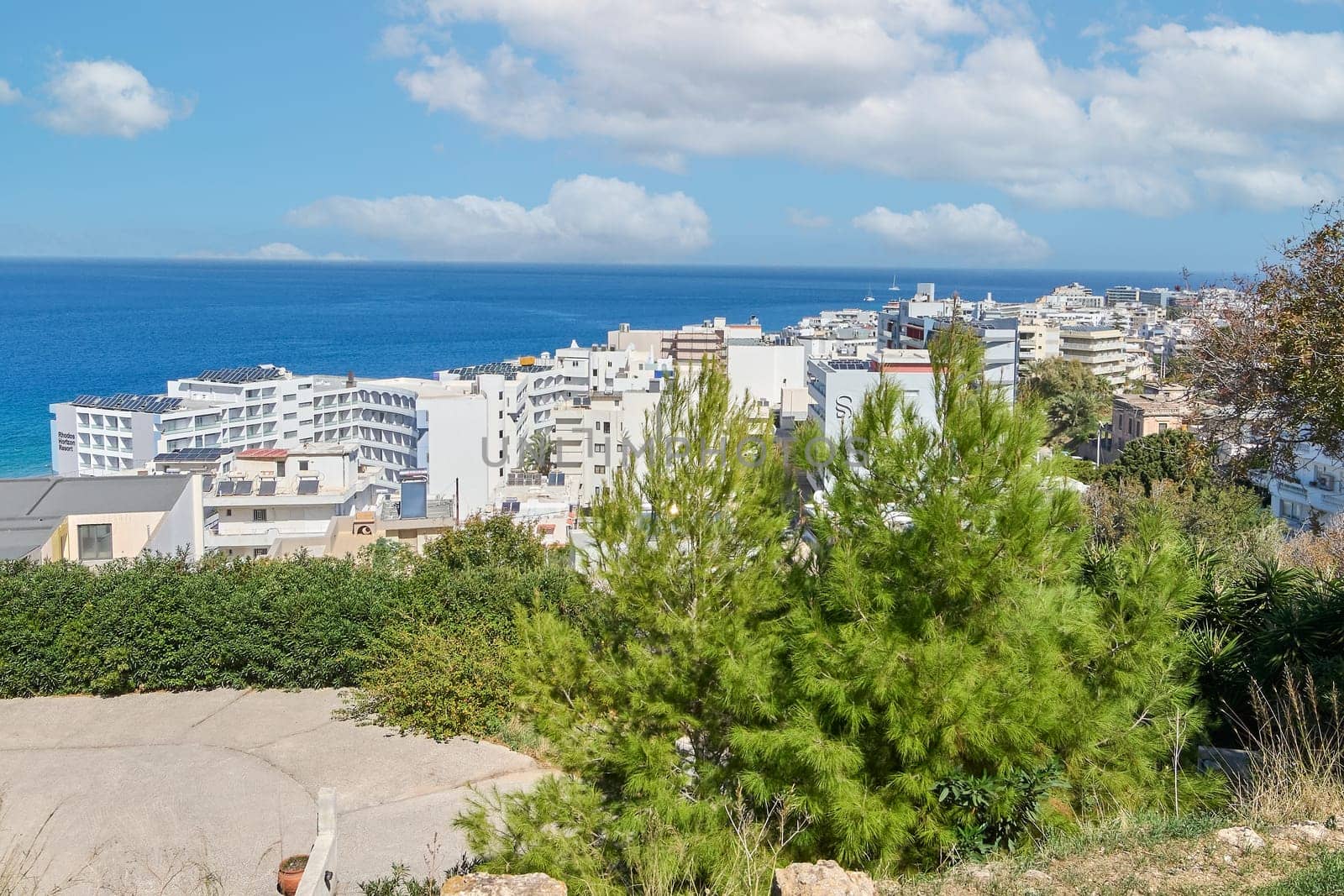 Rodas.Greece - june 07, 2025:  Panoramic view of a vibrant Mediterranean coastal city under a clear blue sky, showcasing white buildings cascading down hillsides towards the sparkling sea